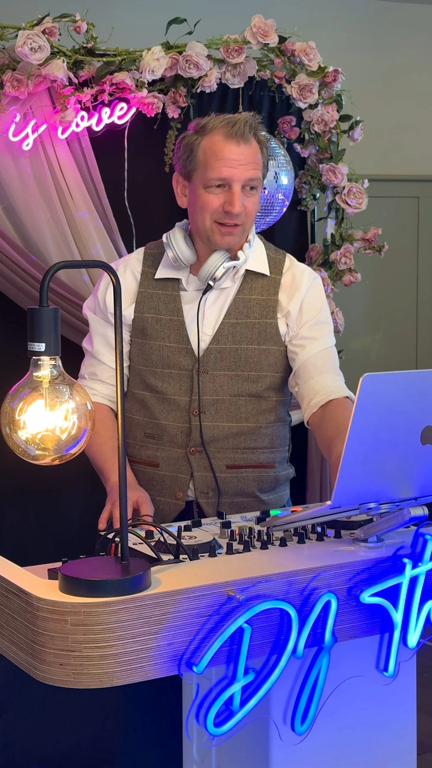 A DJ wearing headphones is standing behind a DJ booth with a laptop, mixing music. The booth has a blue neon sign and is decorated with pink roses, a black curtain, and a disco ball. There is a pink neon sign that says 'love' in the background.