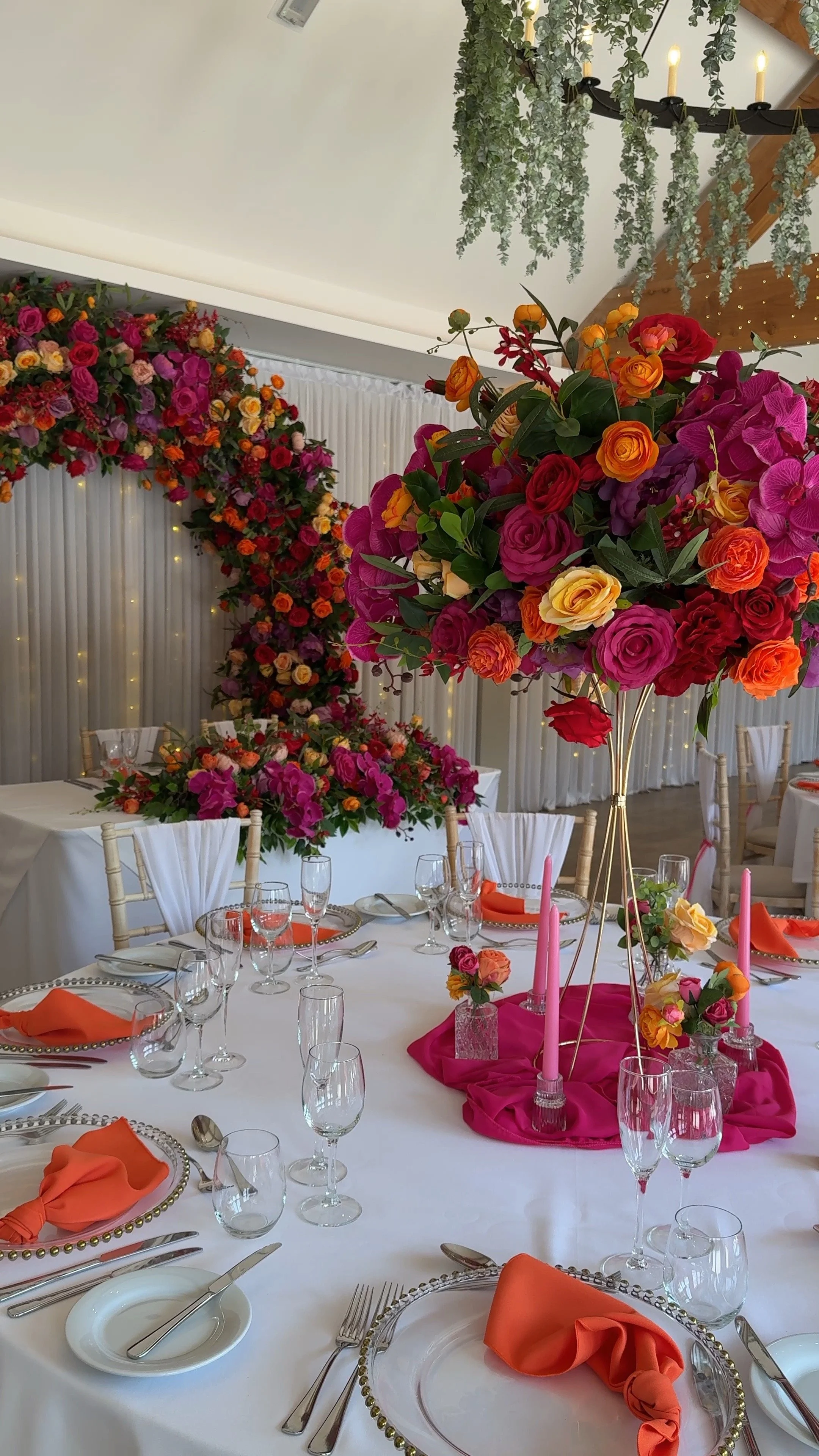 Elegant banquet table decorated with vibrant pink, orange, and yellow flowers, tall pink candles, and orange napkins, set for a celebration in a decorated event space with floral arrangements and hanging greenery.