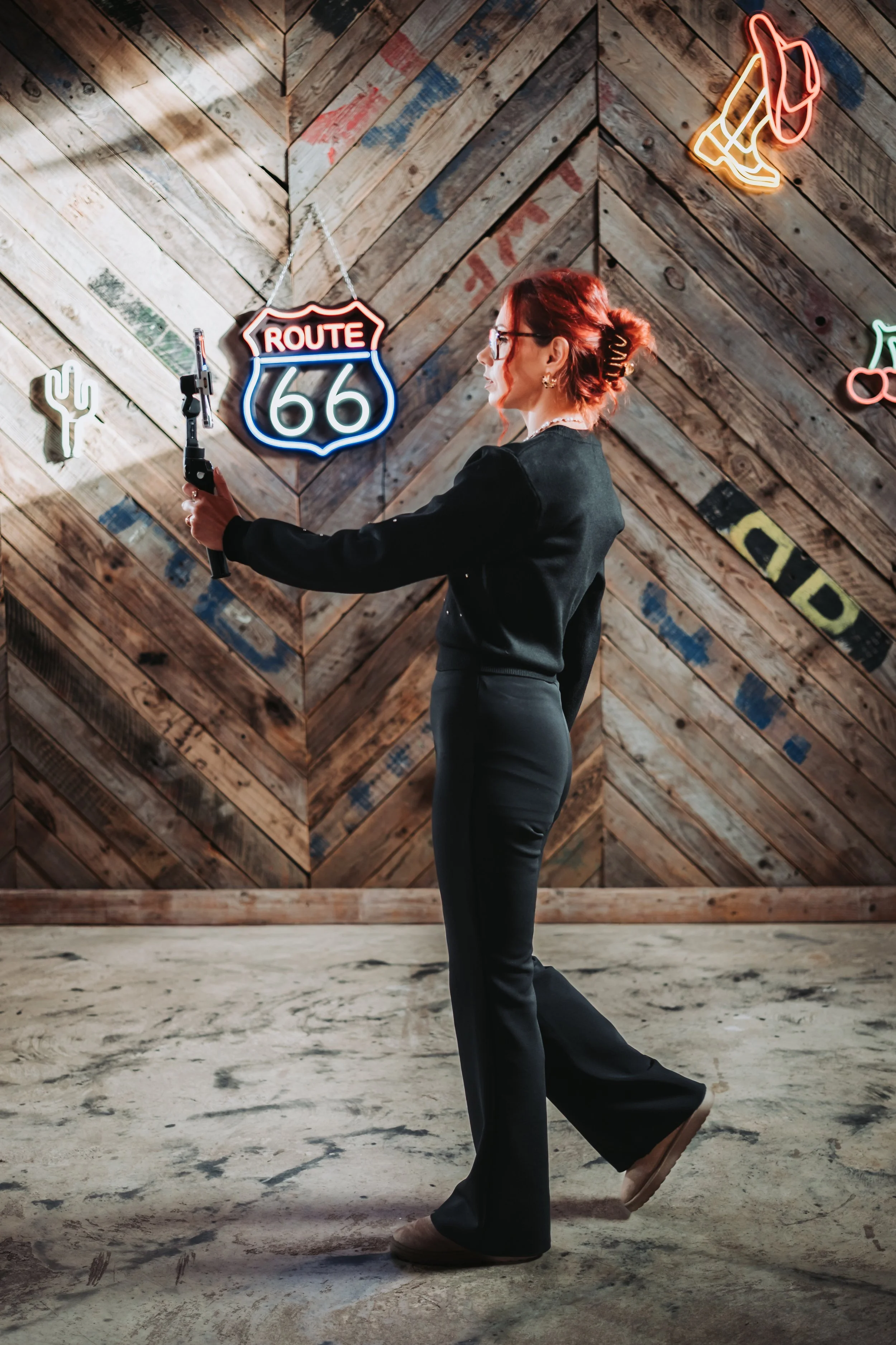 A woman in black pants and a blazer taking a selfie with her phone on a wooden wall background. Neon signs read Route 66, a cactus, a cowboy hat, a boot, and a cowboy hat with a neckscarf are displayed.