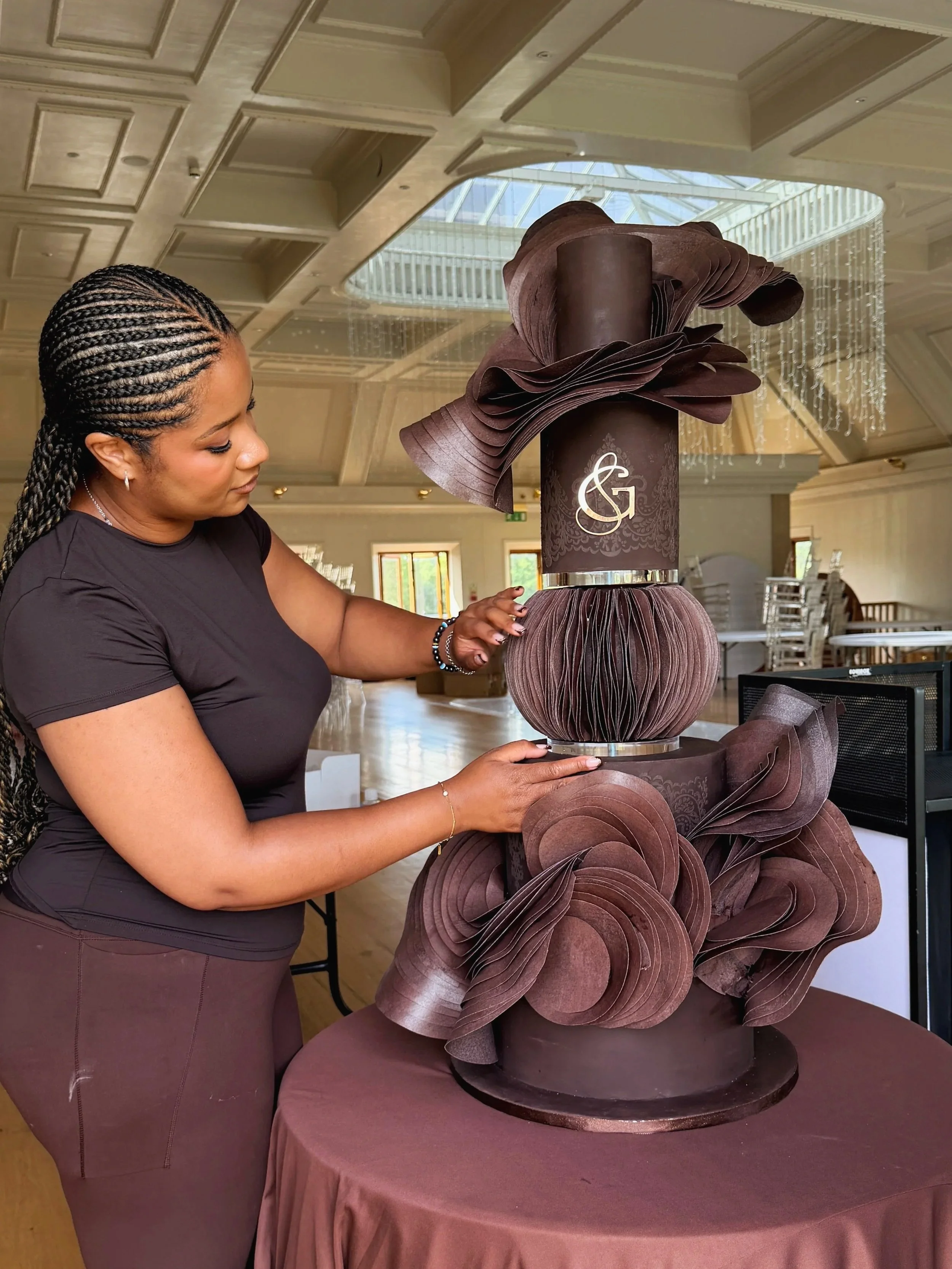 A woman with braided hair, wearing a black top and maroon skirt, is decorating a large, multi-tiered chocolate cake with elaborate, petal-like chocolate decorations in a spacious, well-lit room with chandeliers and large windows.