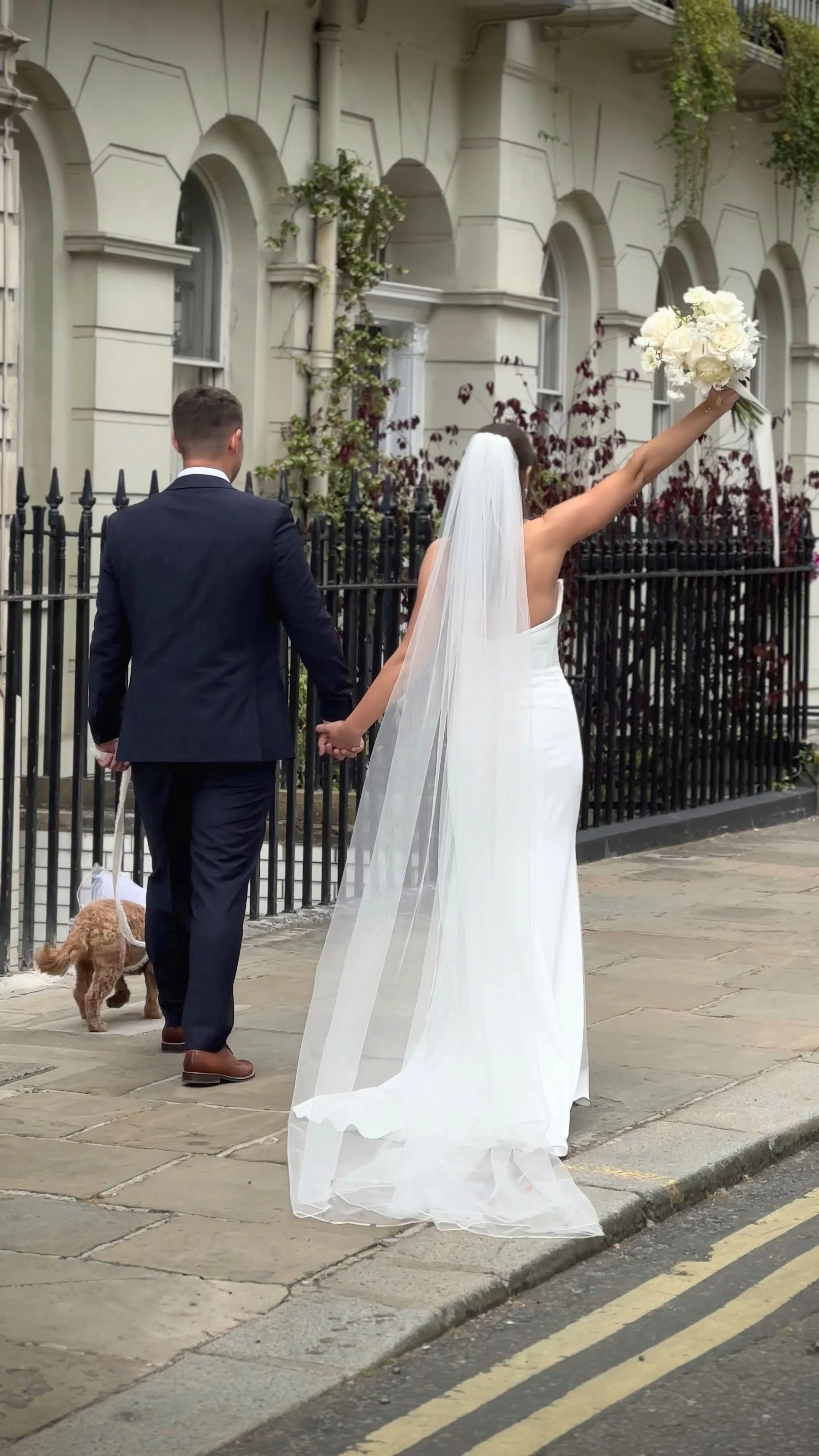 A bride in a white wedding gown and veil, holding a bouquet of white flowers, walking hand-in-hand with a groom in a dark suit while a small dog on a leash walks beside them on a city sidewalk.
