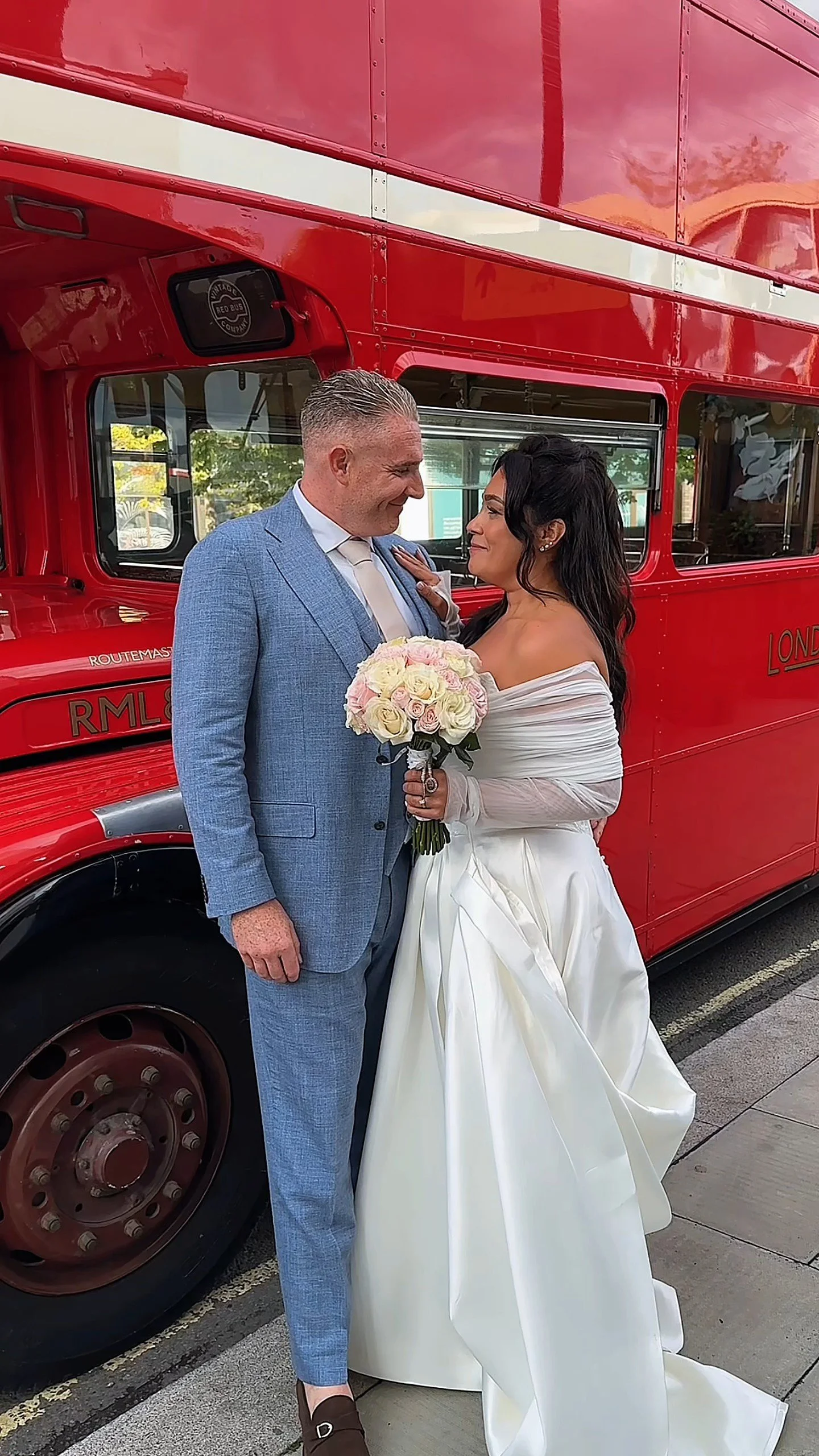 A bride and groom stand close together in front of a red double-decker bus, holding a bouquet of white and pink roses. They are smiling at each other, dressed in wedding attire, with the bride in a white off-shoulder gown and the groom in a light blue suit.
