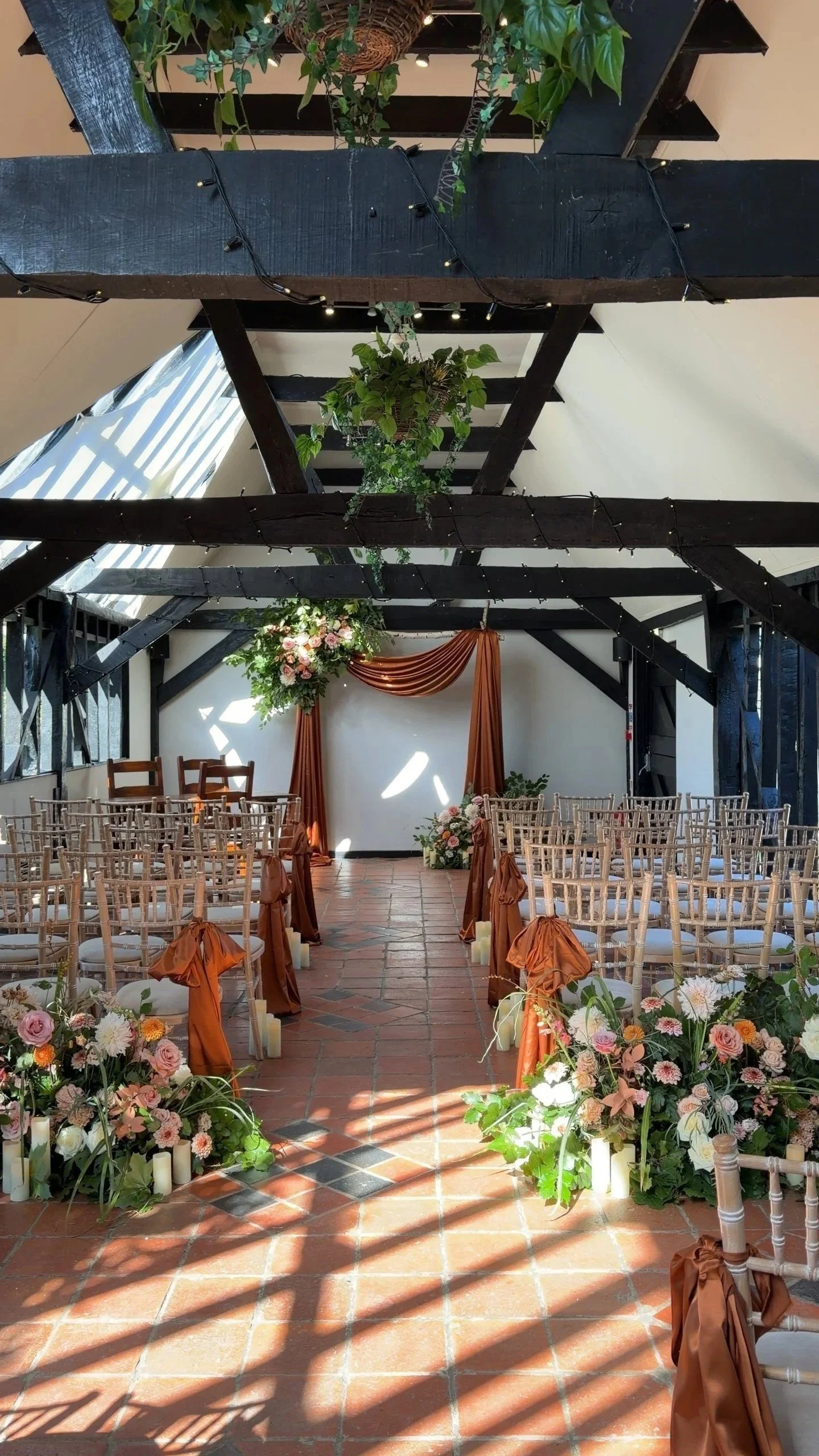 Wedding ceremony setup with chairs facing an altar decorated with floral arrangements and drapes, in an indoor space with expose beams, plants, and string lights.