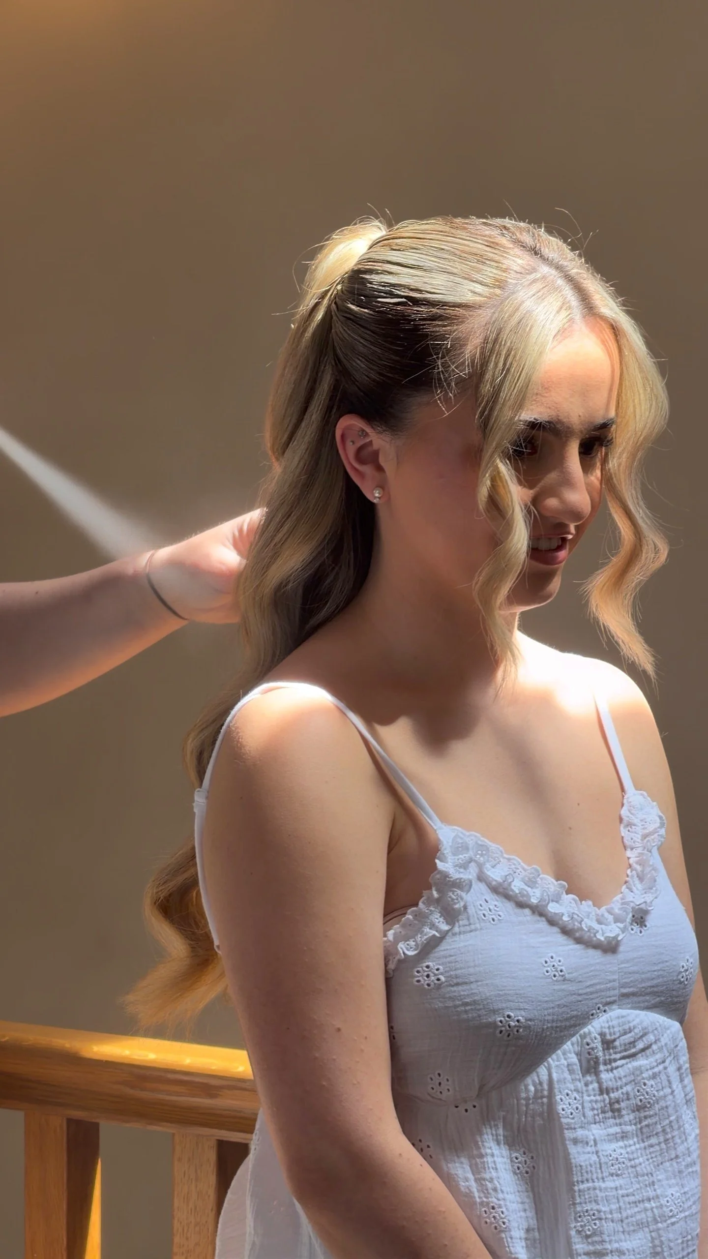 A woman with long, wavy blonde hair wearing a white, sleeveless, lace-trimmed dress sitting on a wooden staircase, while someone styles her hair.