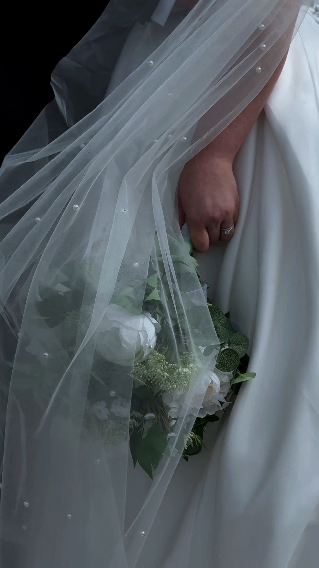 Close-up of a bride's hand holding a bouquet of white flowers, wearing a wedding ring, draped in a sheer veil with pearl accents, and white wedding dress.