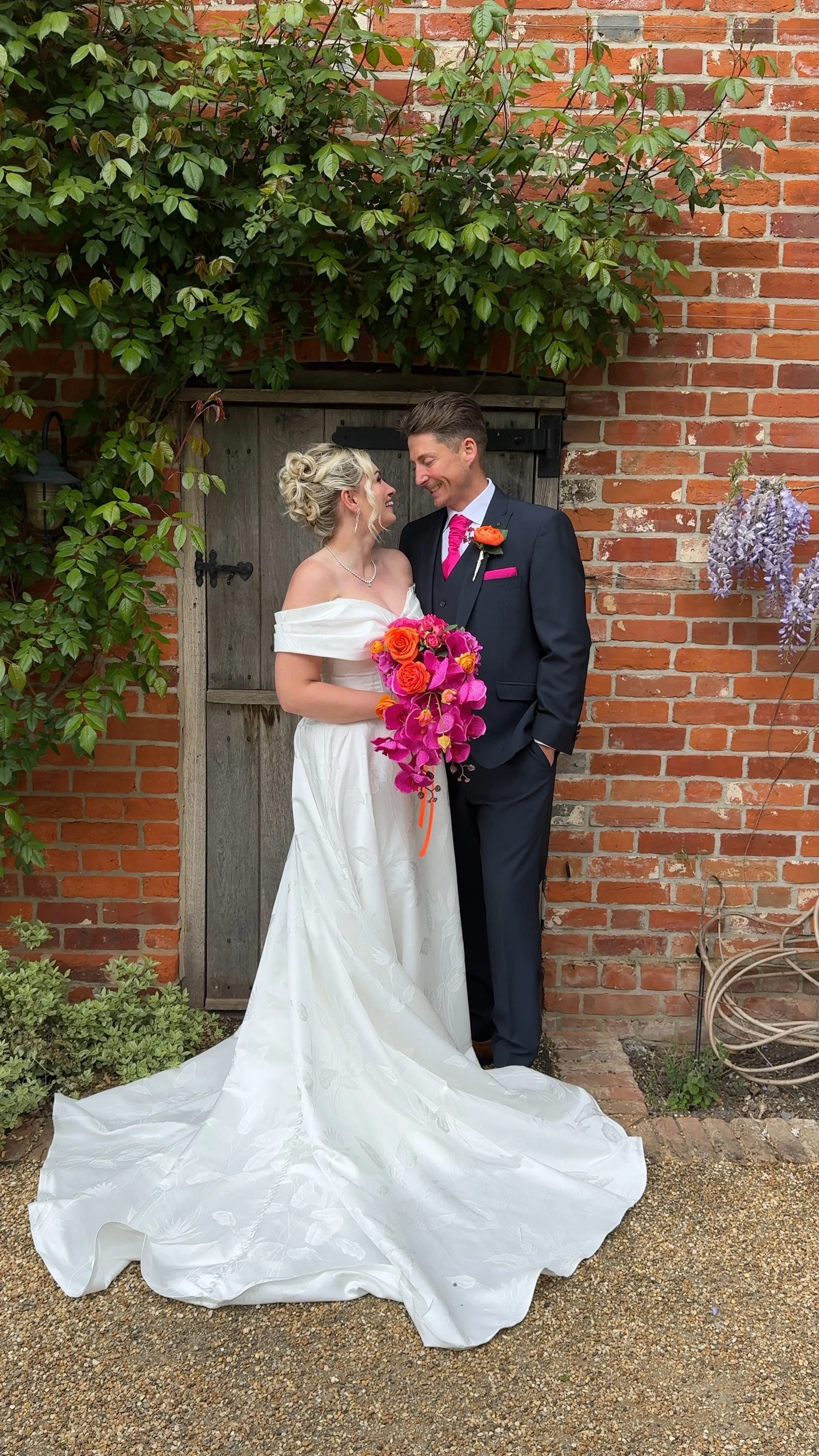 A bride and groom smiling at each other in front of a brick wall with green foliage. The bride is holding a colorful bouquet of pink, orange, and purple flowers, and wearing a white off-shoulder wedding dress. The groom is in a dark suit with a pink tie and orange boutonniere.