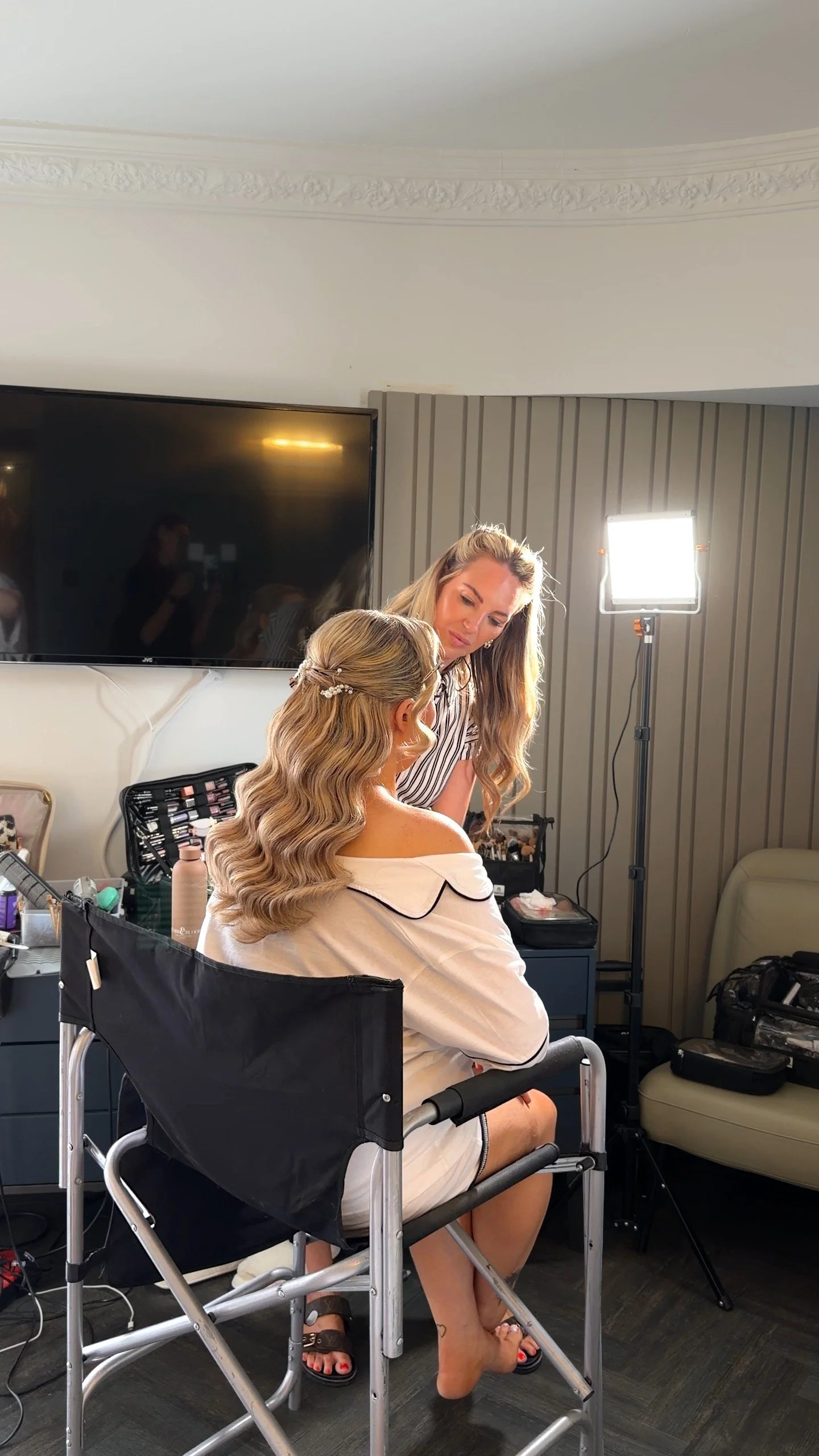 A woman with long black wavy hair and a white off-shoulder top seated in a makeup chair, receiving makeup application from a blonde makeup artist in a striped shirt. The scene is in a room with studio lighting and makeup supplies visible on a table.