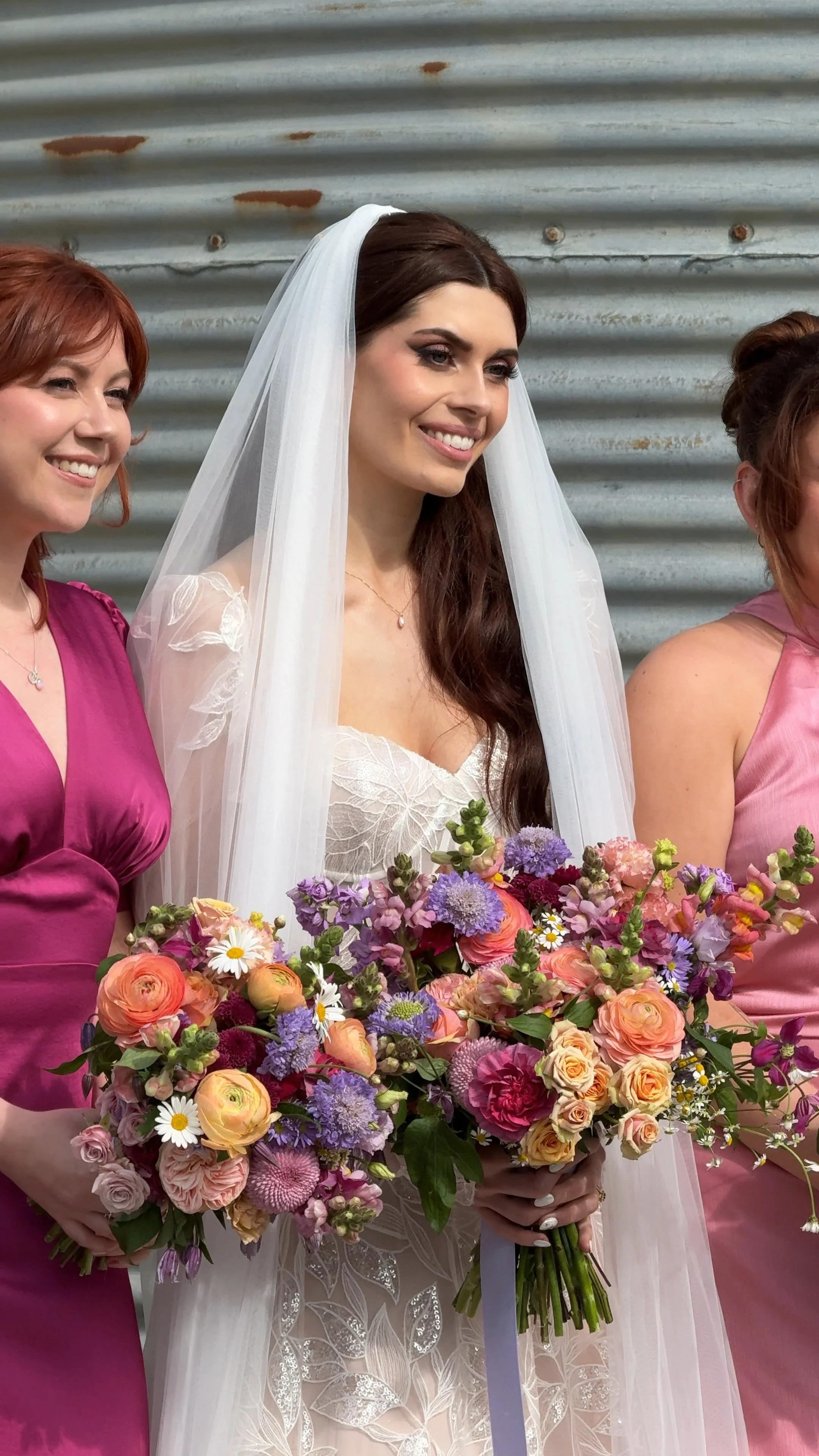 A bride in a white lace wedding dress with a veil, holding a large colorful bouquet, standing next to women in pink dresses against a metal wall background.