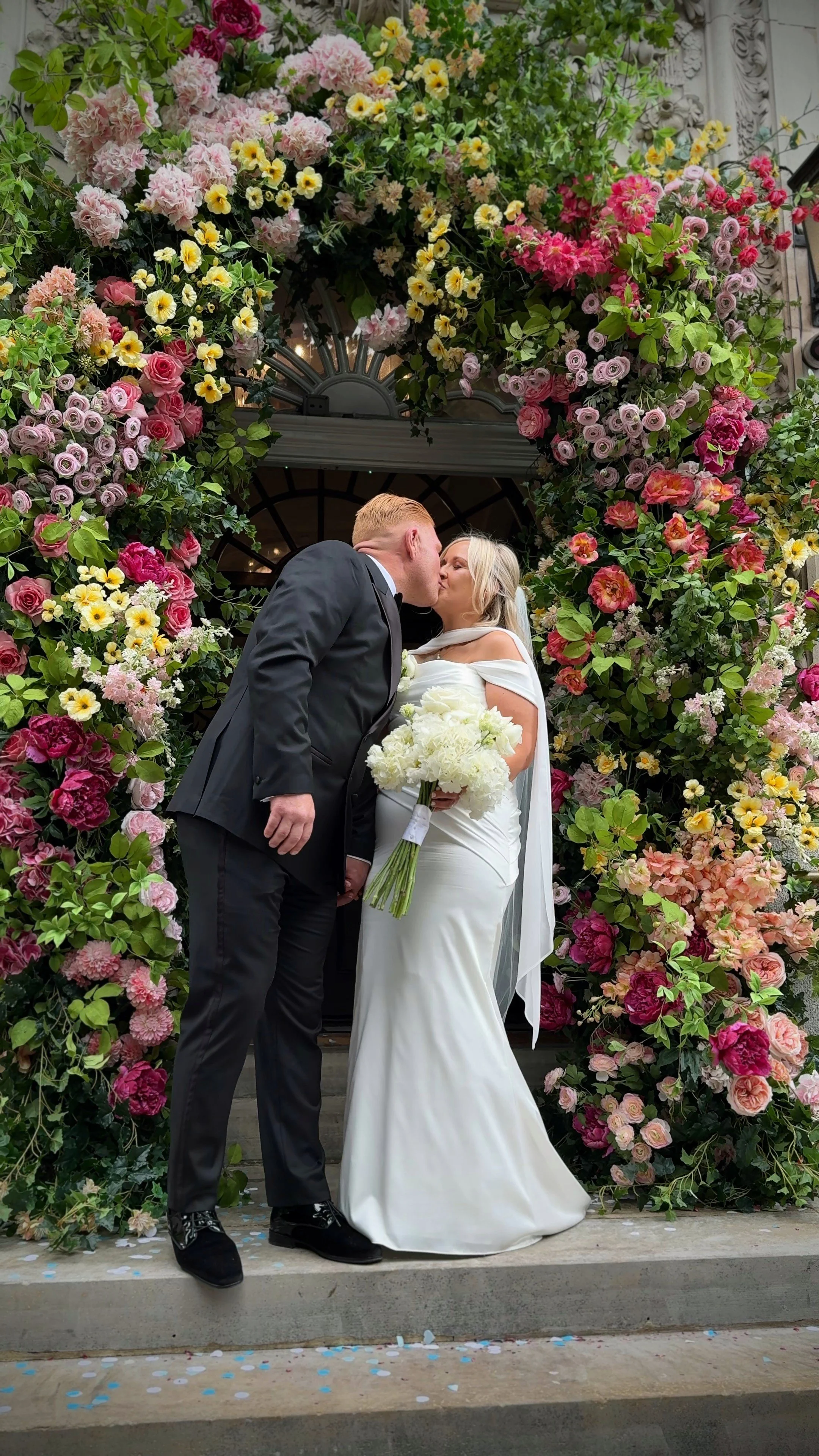 A bride and groom kiss during their wedding ceremony beneath a floral arch with pink, yellow, and purple flowers.