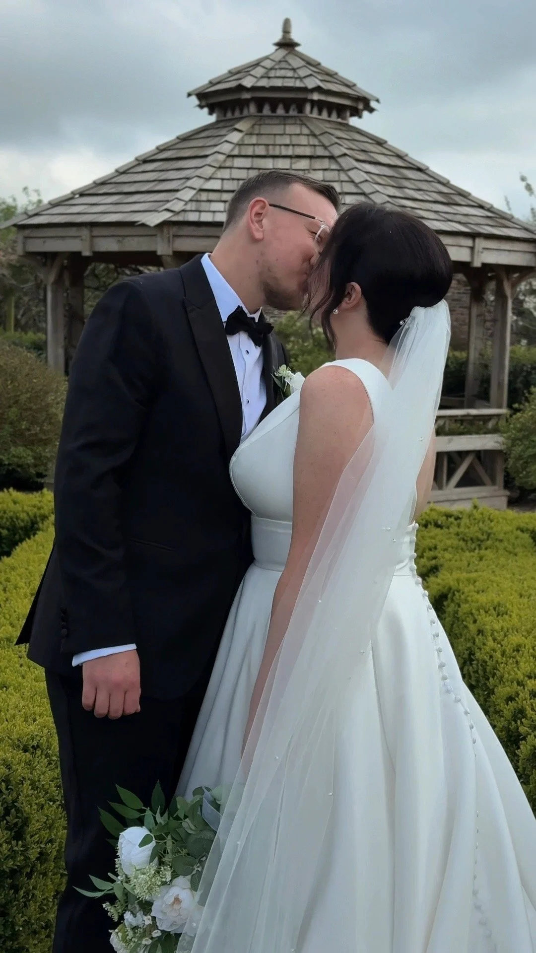 A bride and groom sharing a kiss outdoors in front of a wooden gazebo, surrounded by greenery.