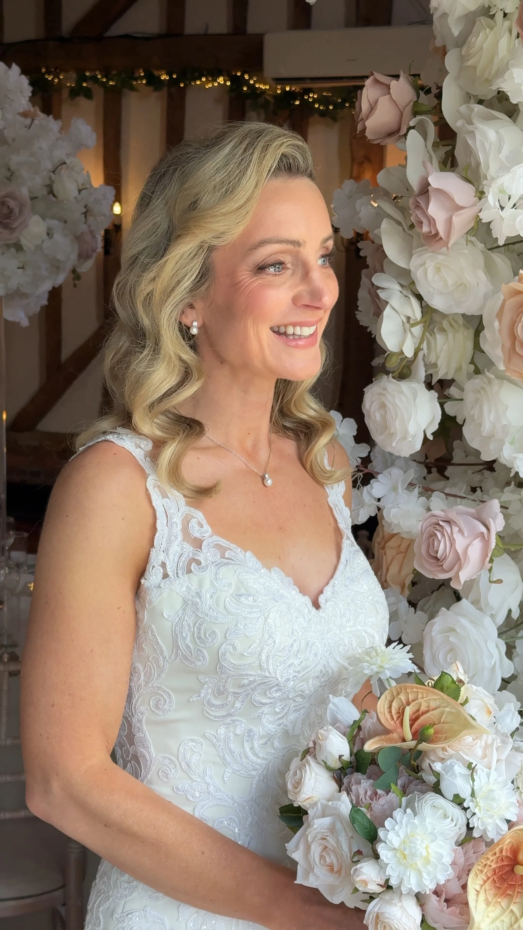 A woman in a white lace wedding dress standing near a floral arrangement of white, blush, and peach roses and calla lilies, indoors with wooden beams and soft lighting.
