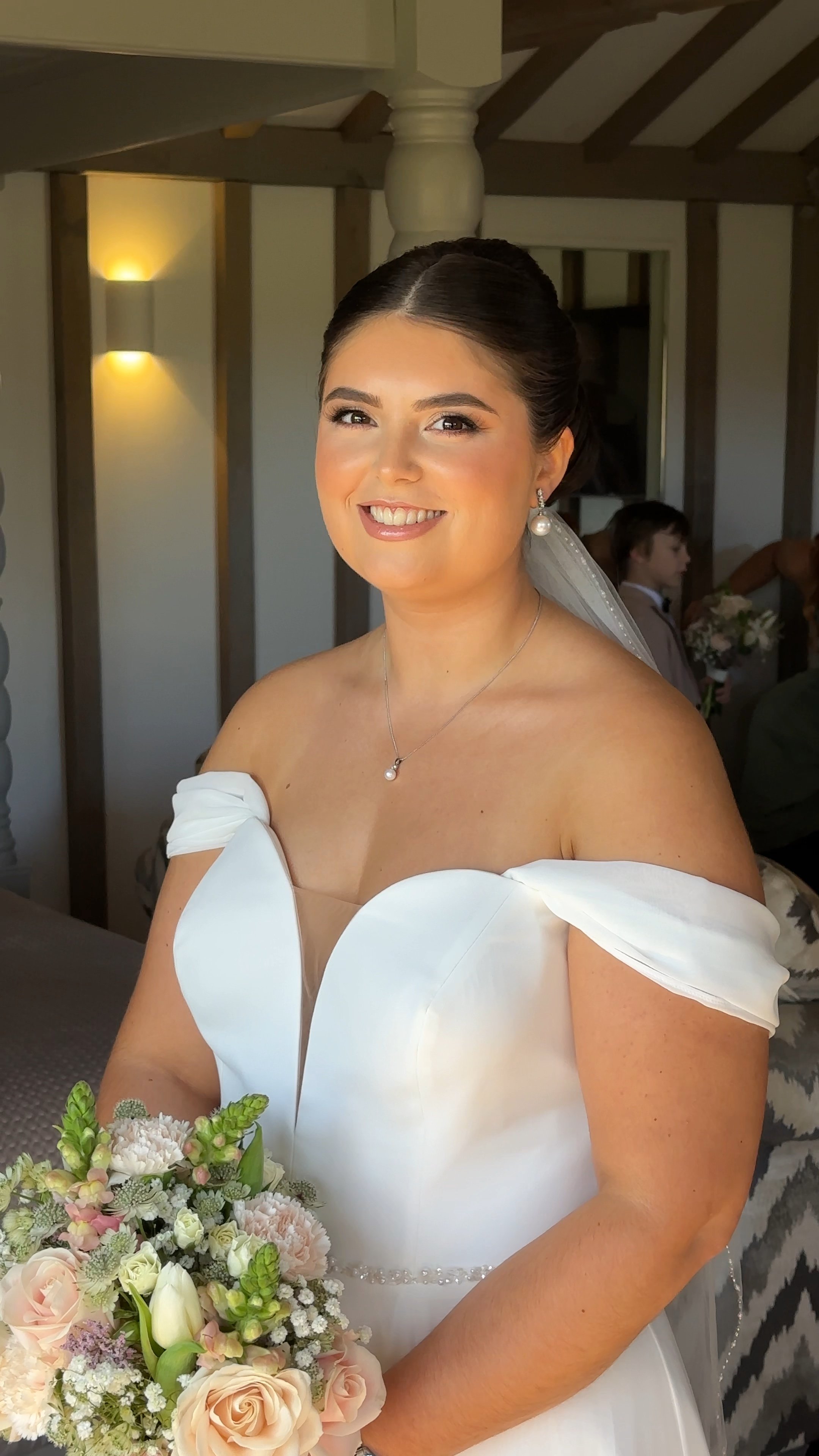 A smiling bride wearing a white off-the-shoulder wedding dress, pearl earrings, necklace, and veil, holding a bouquet of pale pink and white flowers. The background shows warmly lit walls and a young boy holding a bouquet in a room with wooden beams.