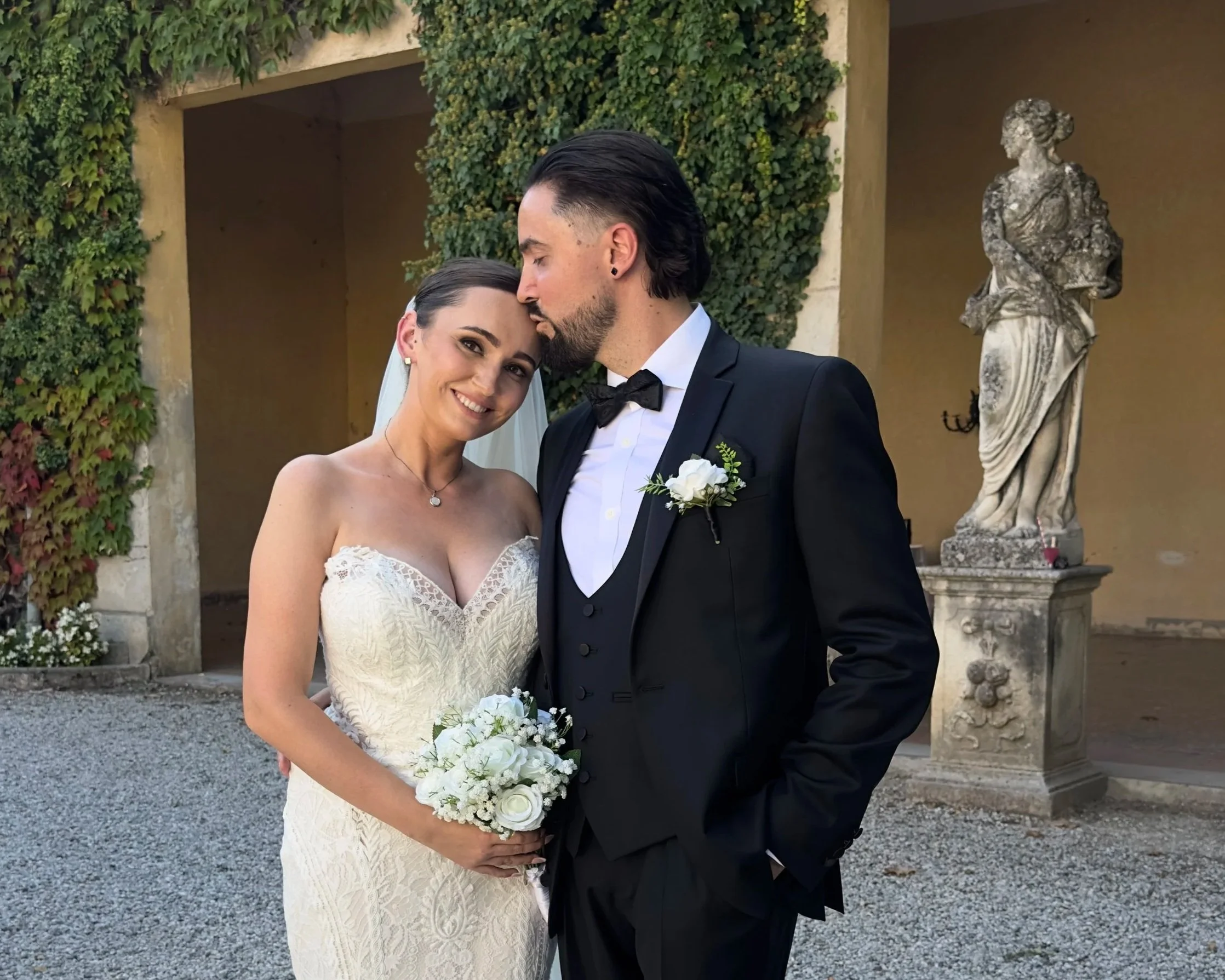 A bride and groom embrace on their wedding day outdoors near a classical statue and a wall covered with climbing plants. The bride wears a strapless white lace wedding gown and holds a bouquet of white roses. The groom wears a black tuxedo with a bow tie.