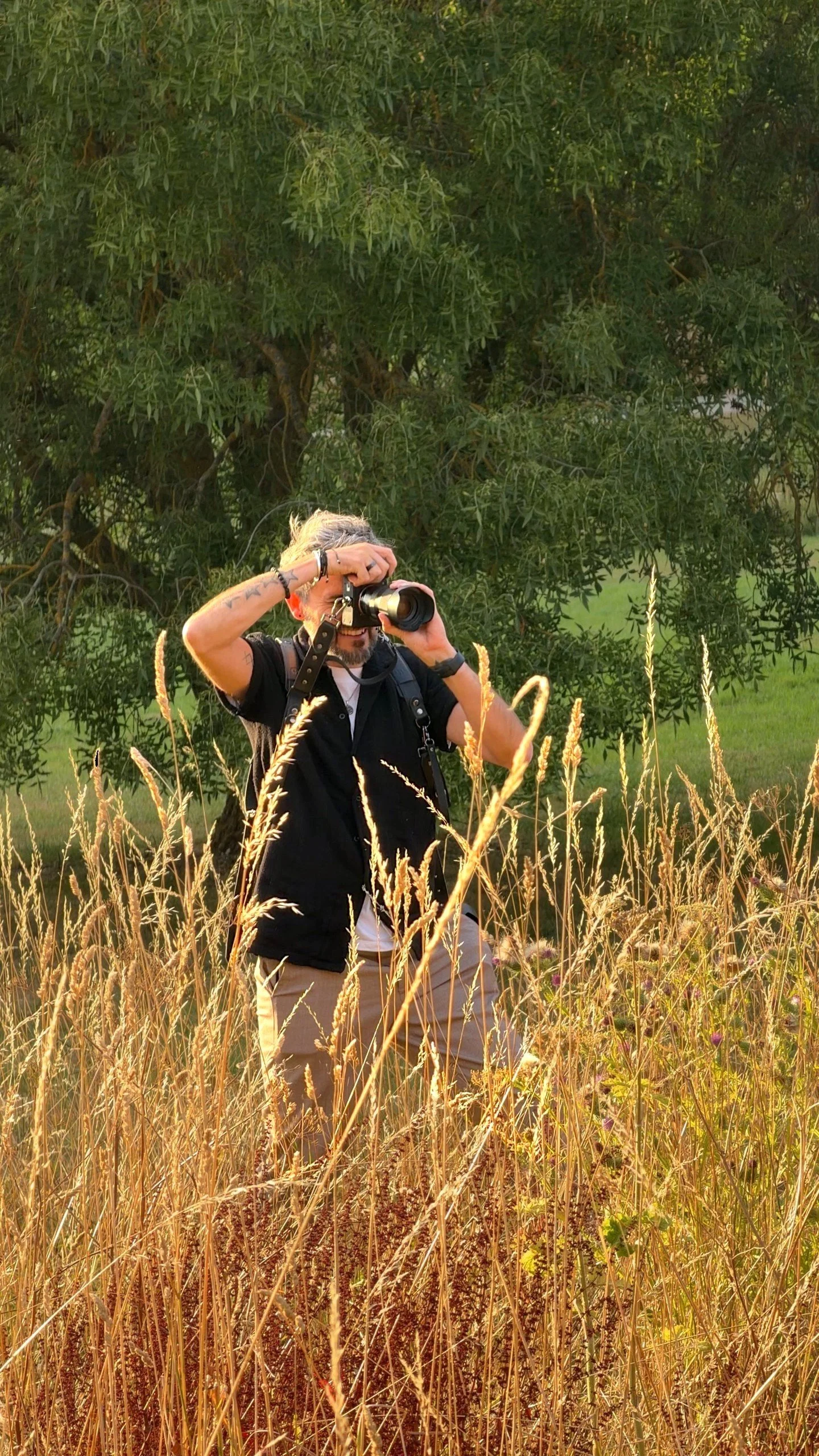 A man with gray hair and tattoos taking a photograph with a camera in a grassy field, with green trees in the background.