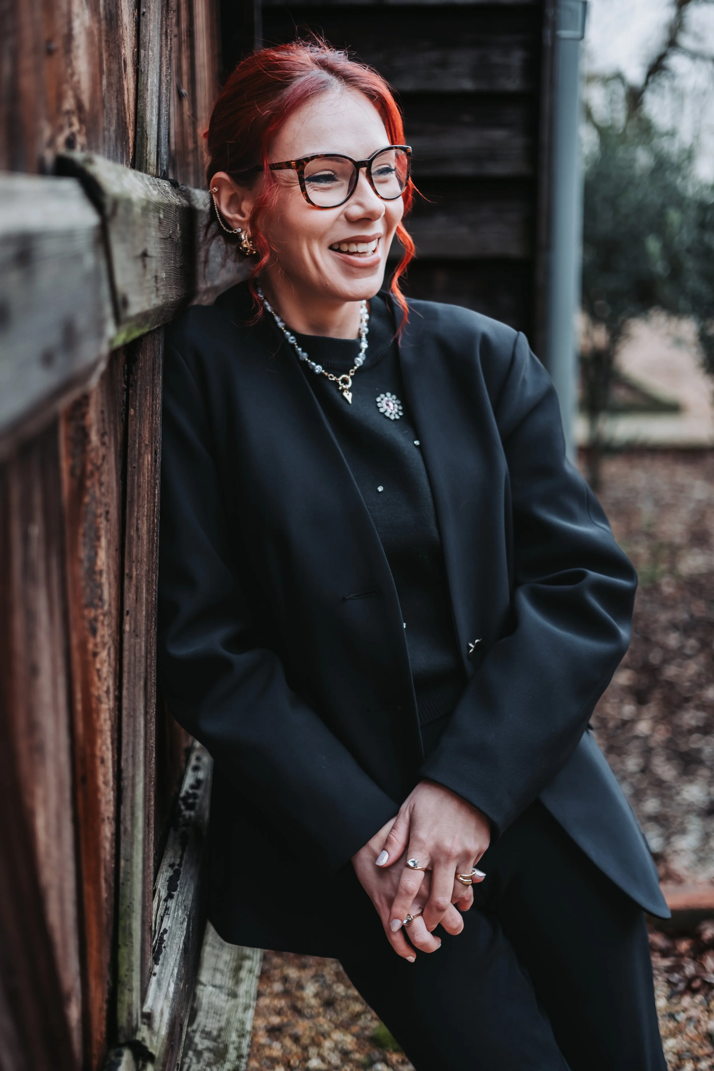 A woman with red hair and glasses leaning against a weathered wooden fence outdoors, smiling and wearing a black jacket with jewelry, with trees blurred in the background.