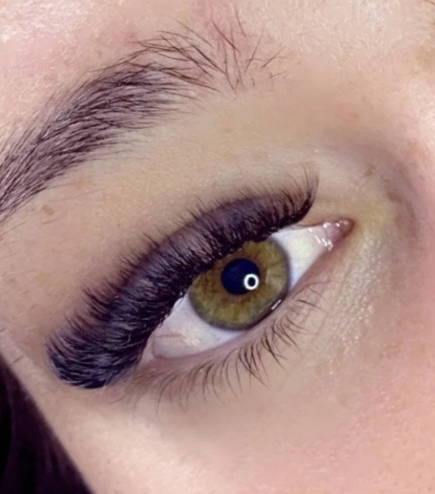 Close-up of a person's eye with greenish-brown iris, featuring long, beautiful signature style of eyelash extensions and well-groomed eyebrows