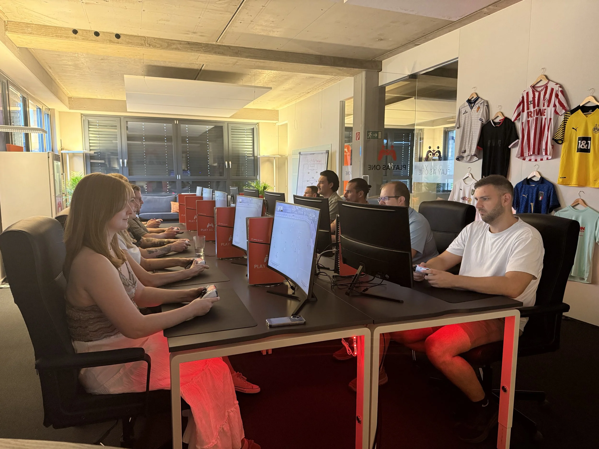Menschen sitzen in einem Büro vor Computern. Es gibt Sporttrikots an der Wand und einen Whiteboard-Tafel.