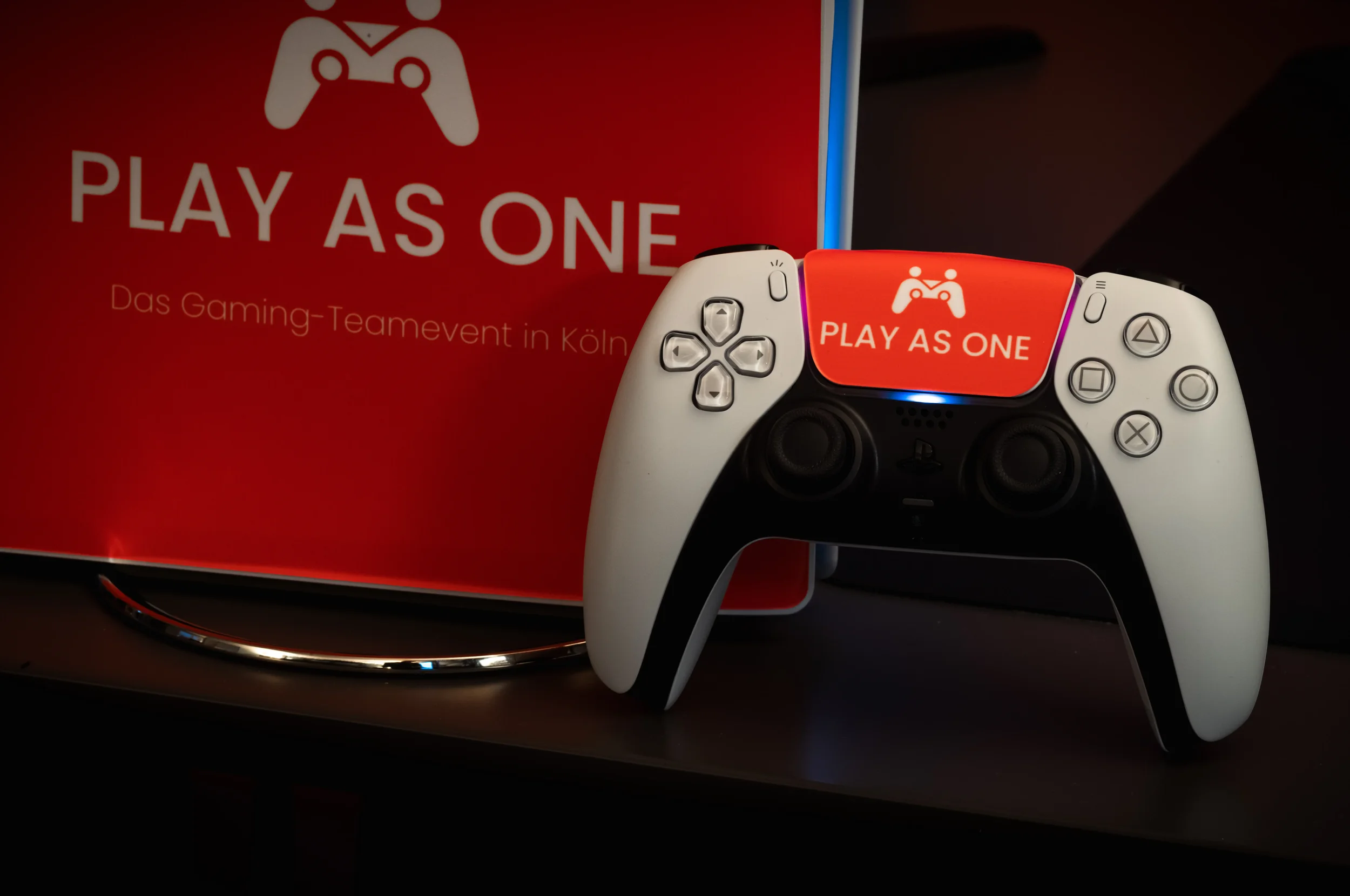 Ein PlayStation-Controller mit einem Aufkleber mit der Aufschrift 'PLAY AS ONE' und einem Spiele-Logo, neben einem Bildschirm, auf dem ebenfalls die gleiche Aufschrift zu sehen ist, in einem dunklen Raum.