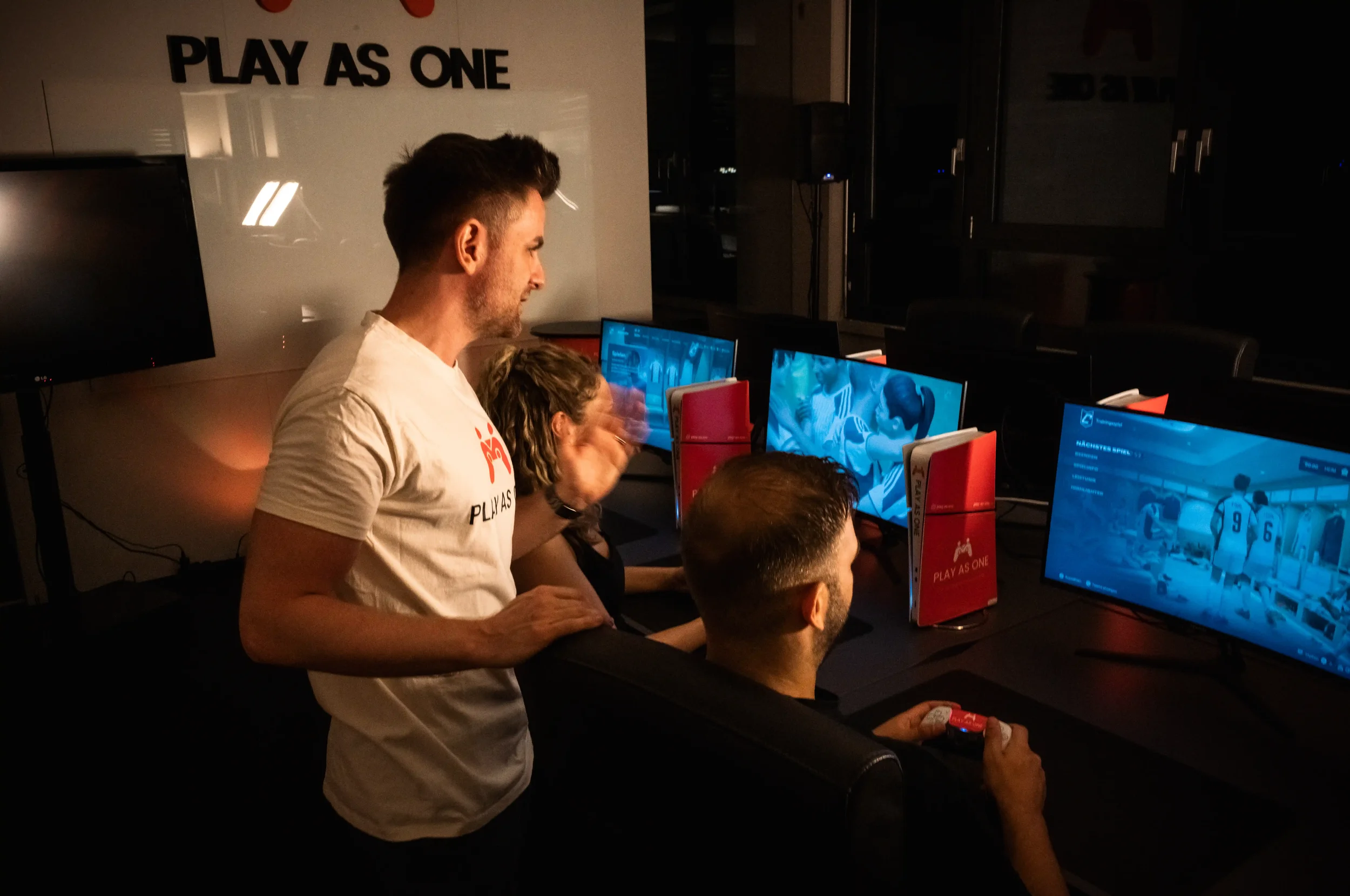 Mehrere Menschen spielen an Computern, ein Mann steht und beobachtet die Bildschirme, im Hintergrund ein Whiteboard mit dem Text 'PLAY AS ONE'.