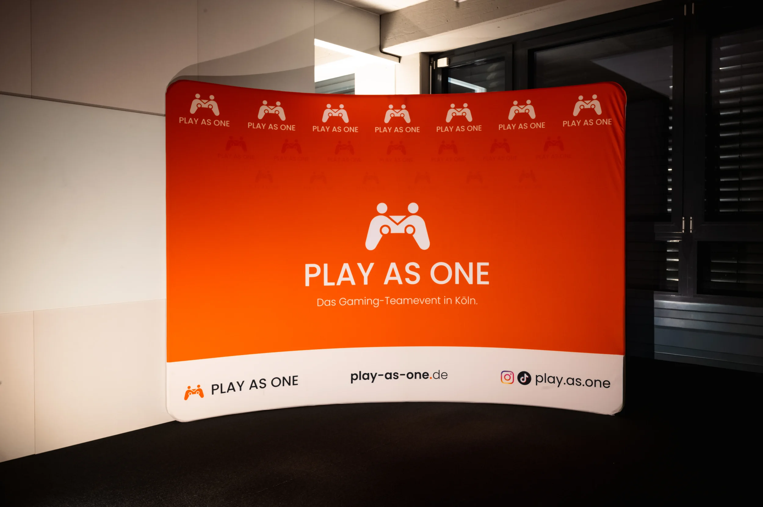 Ein rotes Banner mit einem Spiele-Controller-Symbol und dem Text "PLAY AS ONE" sowie weiteren wiederholten Symbolen. Unten steht "Das Gaming-Teamevent in Köln". Das Banner enthält auch Web- und Social-Media-Links.