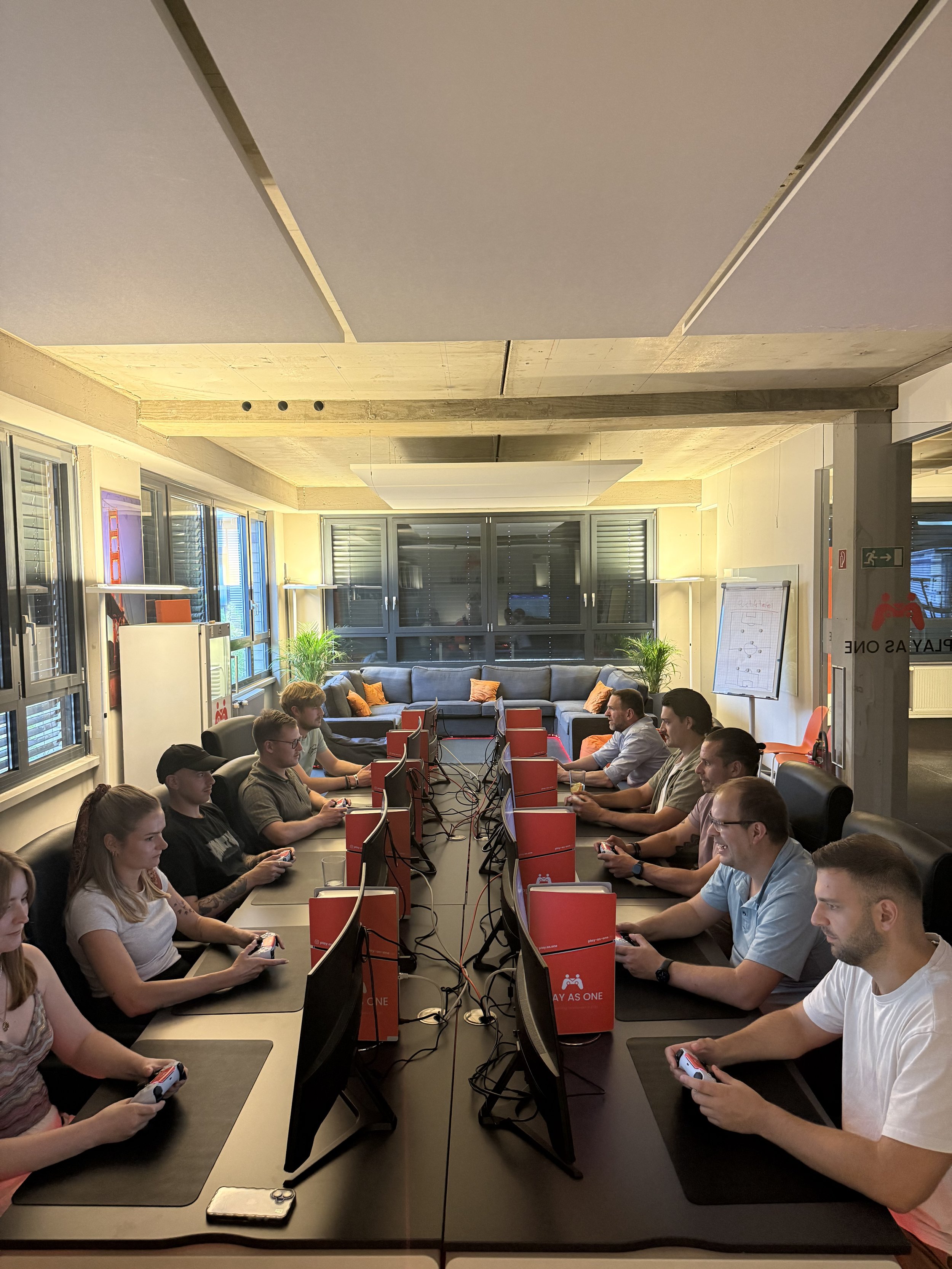 Ein Raum mit mehreren Menschen, die an einem Tisch mit Computern und Spielkonsolen sitzen, vermutlich bei einem Spiele-Event oder Turnier.