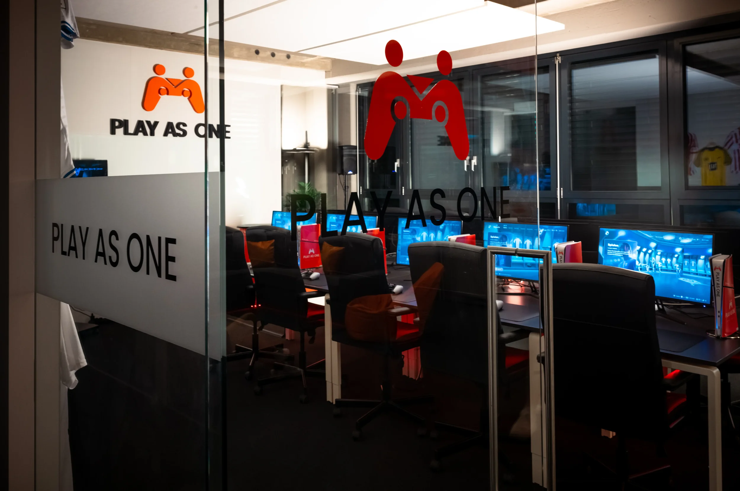 Ein Raum mit mehreren Computern und Stühlen. Im Fenster sind Symbole eines roten Spielers und des Textes 'PLAY AS ONE' zu sehen.