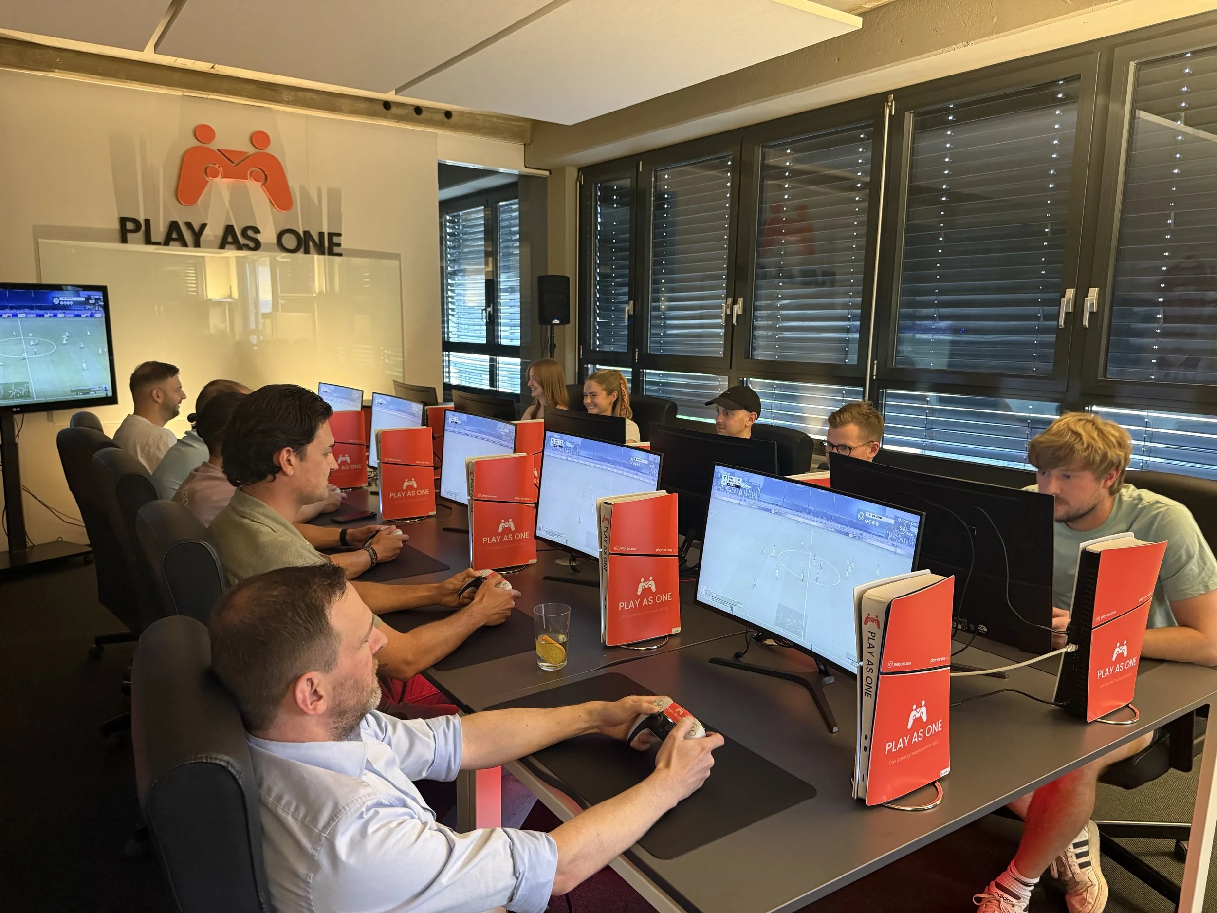 Gruppen von Menschen spielen an Playstations in einem Spielraum, mit einem Poster 'PLAY AS ONE' an der Wand.