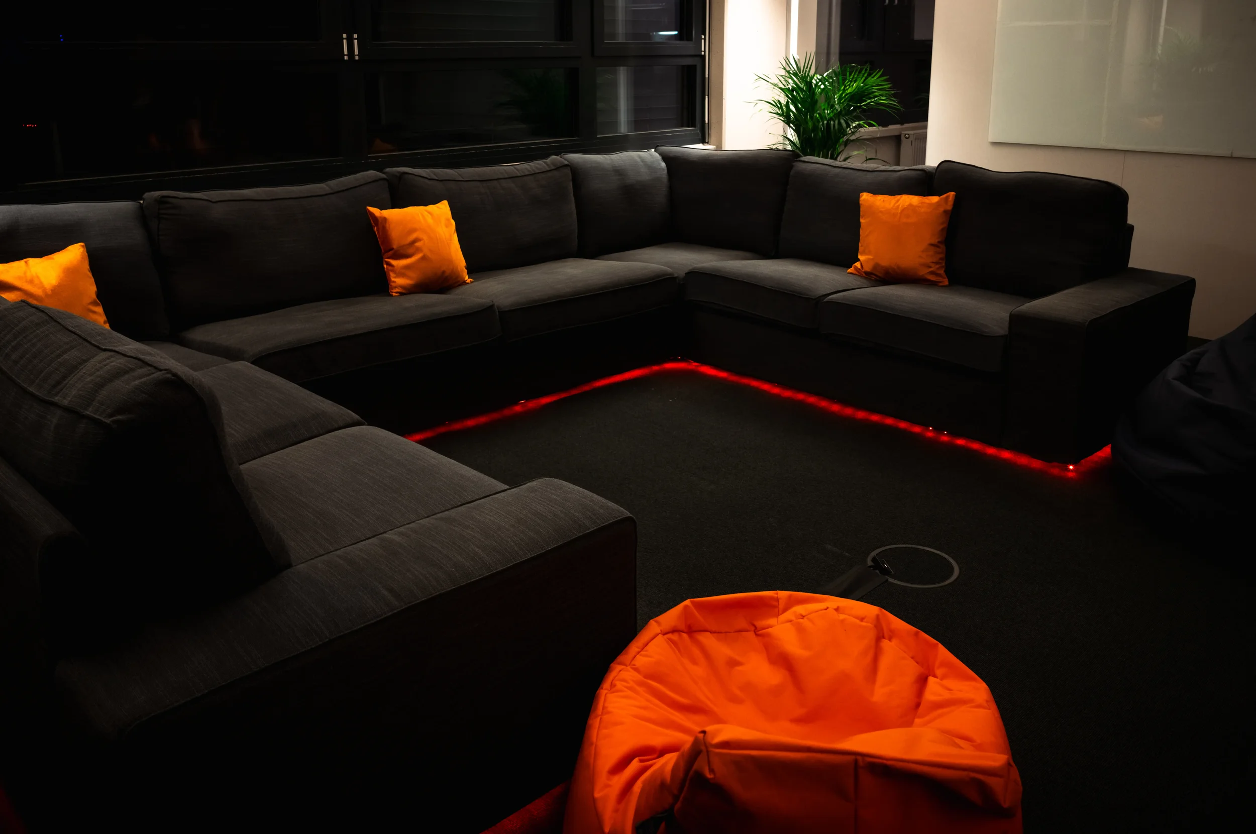 Ein Wohnzimmer mit einer schwarzen Couch, mit orangefarbenen Kissen, im Hintergrund eine große Pflanze und ein Fenster, auf dem Boden eine mit roten Lichtern beleuchtete Linie.