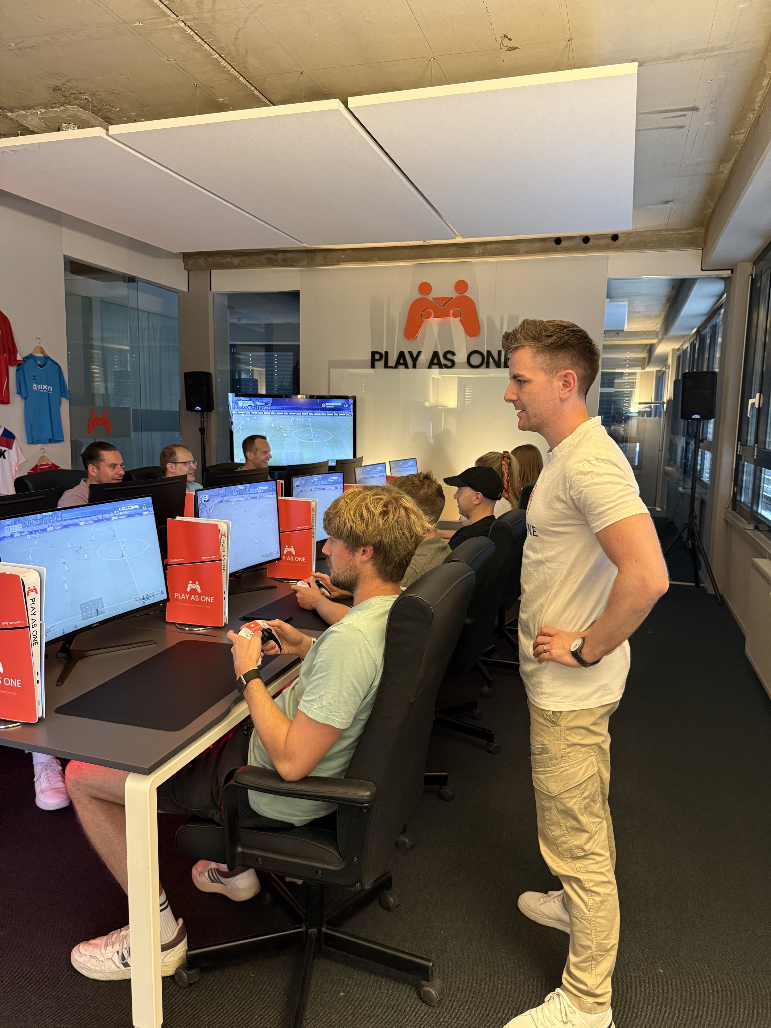 Junge Männer spielen ein Fußball-Videospiel an Computern in einem Raum mit mehreren Monitoren und einem großen Bildschirm, auf dem das Spiel angezeigt wird.