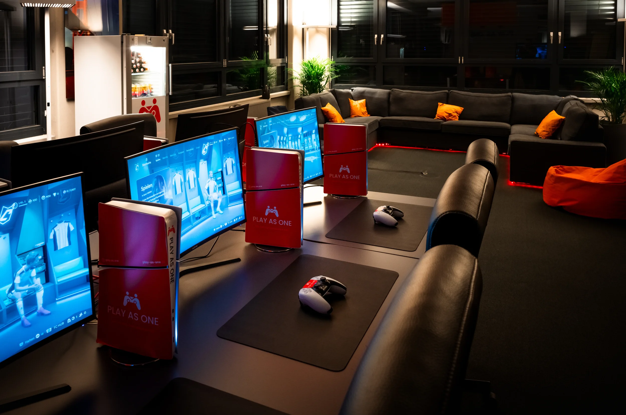 Ein Raum mit mehreren Computerbildschirmen, einer Sofagarnitur mit orangefarbenen Kissen und Pflanzen, vermutlich ein Gaming- oder Aufenthaltsraum.