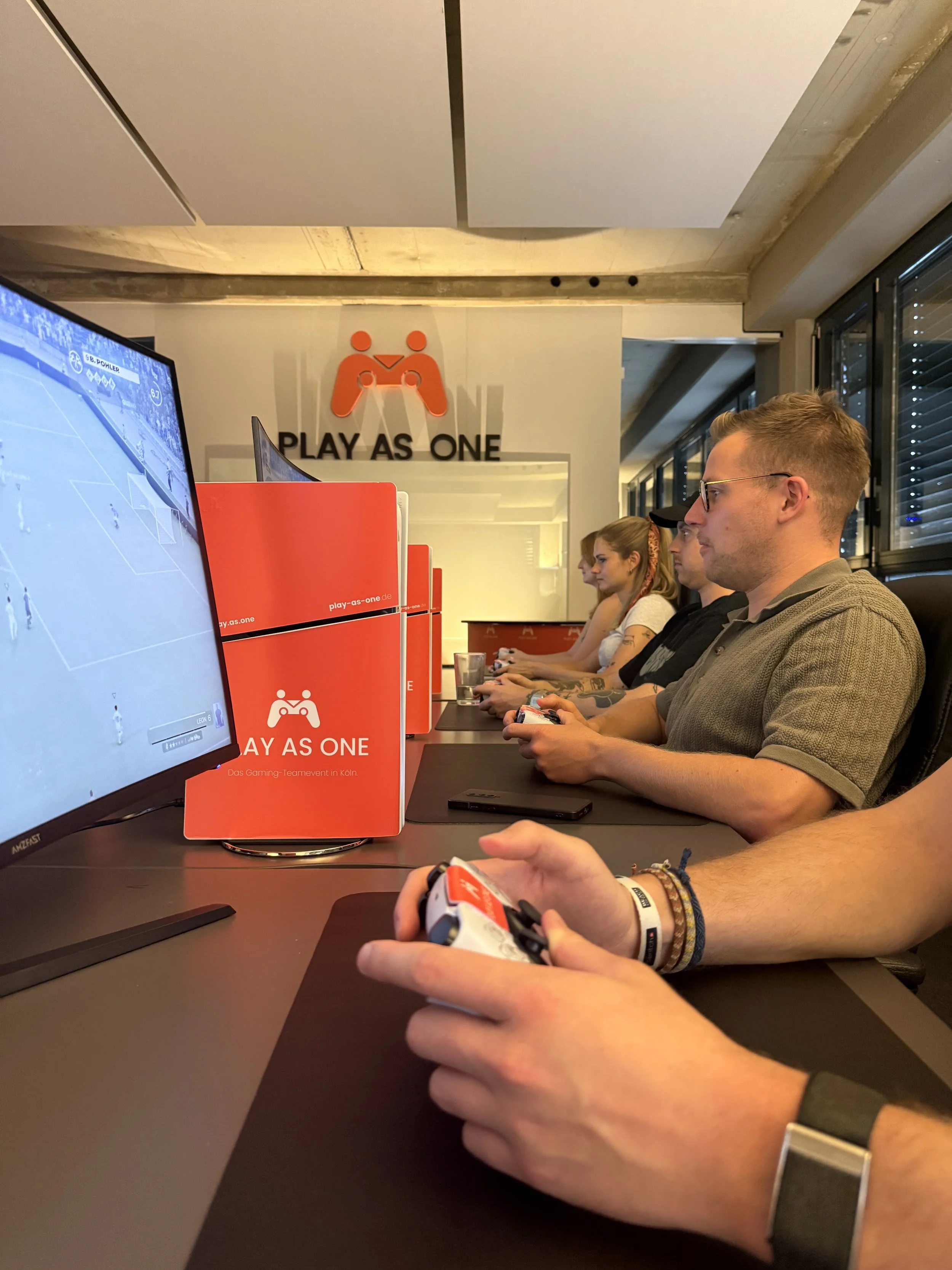 Mehrere Menschen spielen Videospiele an Tischen in einem Raum mit Spielkonzept 'Play as One', auf einem Monitor ist ein Fußballspiel zu sehen, im Hintergrund eine Wand mit einem Logo und dem Schriftzug 'PLAY AS ONE'.