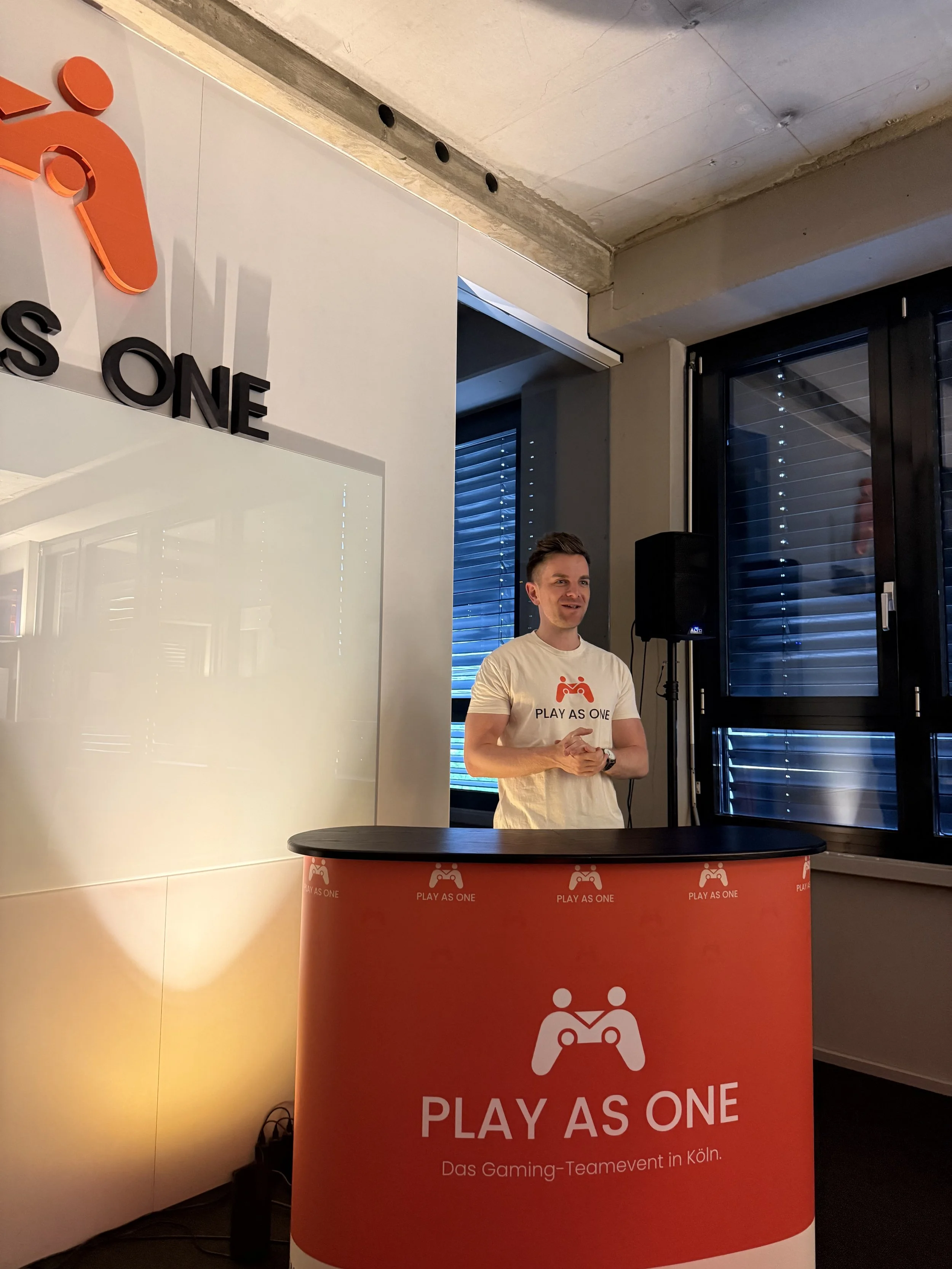 Junger Mann steht hinter einem runden roten Infostand mit dem Logo und der Aufschrift 'PLAY AS ONE' bei einem Gaming-Teamevent in Köln. Im Hintergrund sind Fenster mit geschlossenen Jalousien und eine schwarze Lautsprecherbox zu sehen.