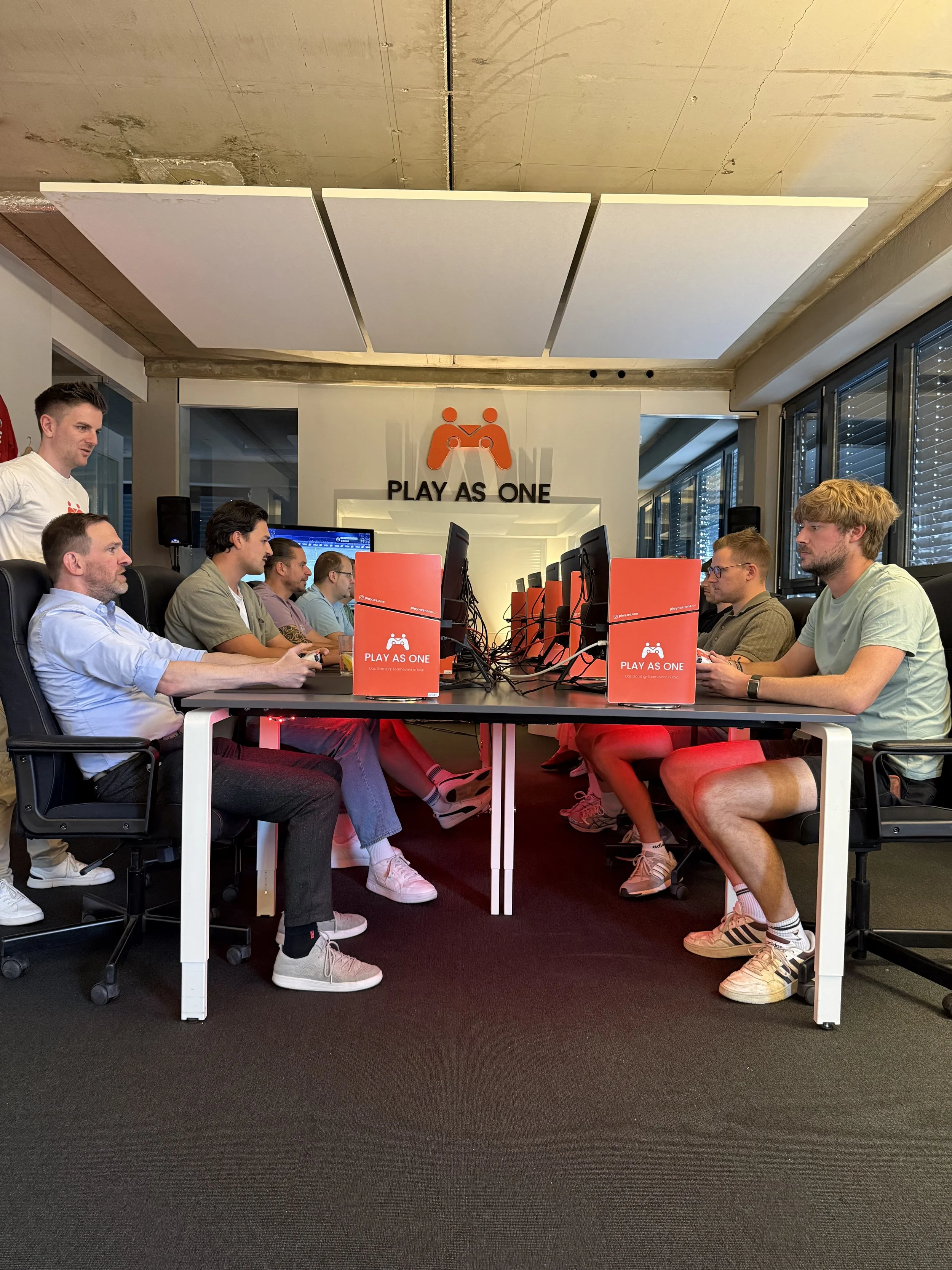 Mehrere Menschen sitzen im Raum vor Computern und spielen ein Spiel. Es ist eine Gaming- oder E-Sport-Veranstaltung mit dem Slogan 'PLAY AS ONE' an der Wand im Hintergrund.