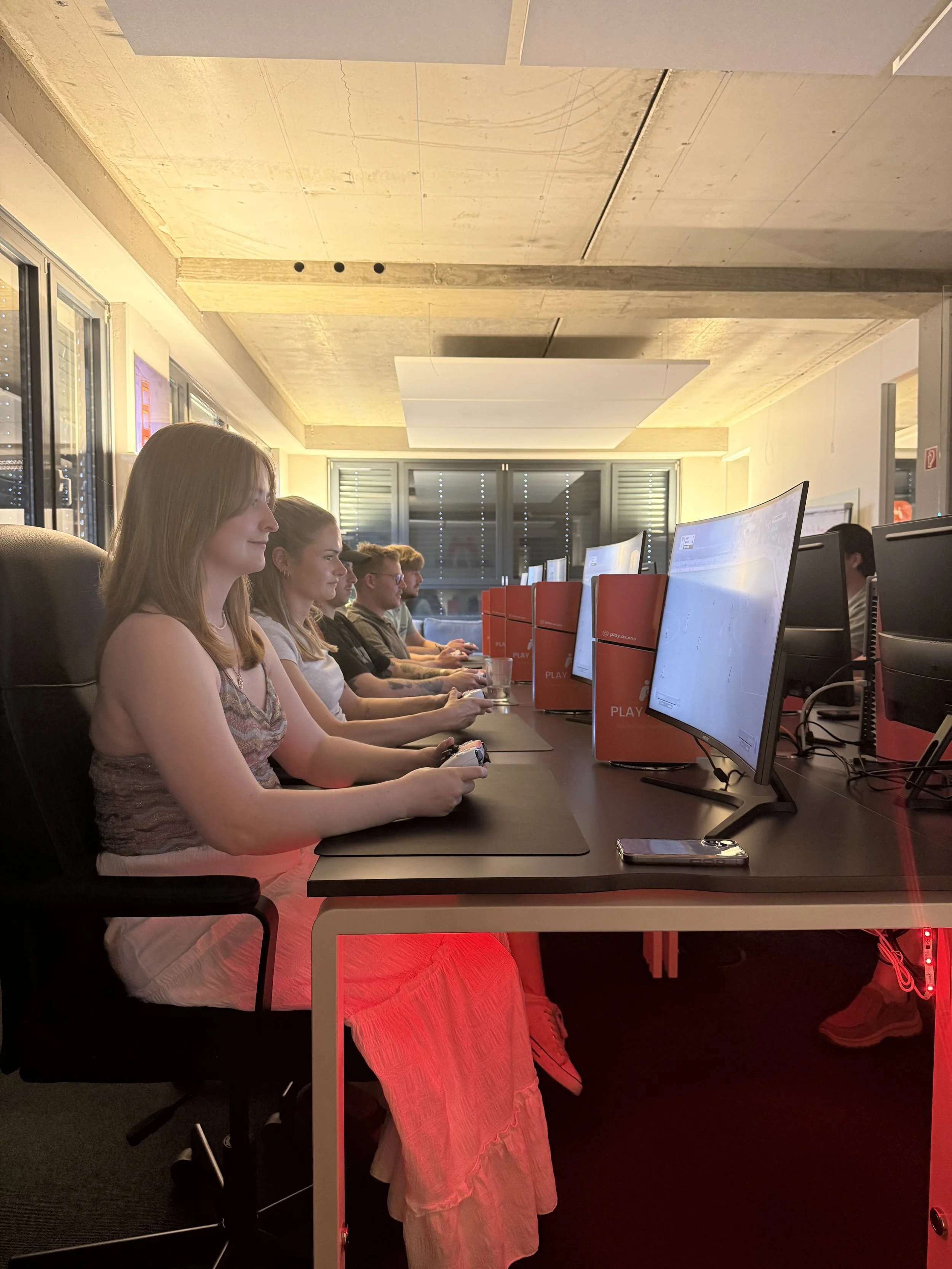Menschen sitzen vor Computern in einem Raum, nutzen Tastaturen und Mäuse, während sie auf ihre Bildschirme schauen, im Hintergrund Fenster mit geschlossenen Jalousien.