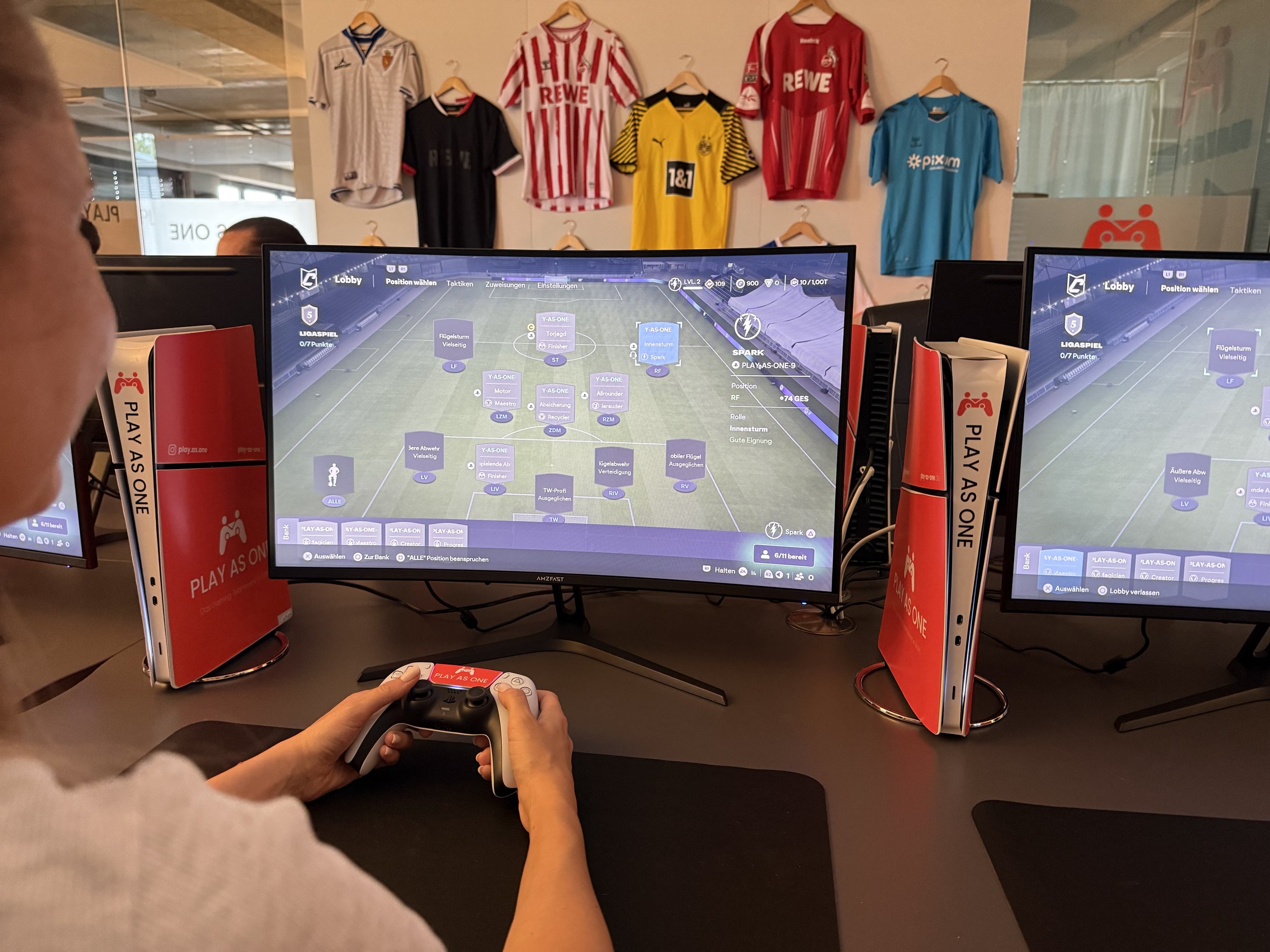 Zwei Computerbildschirme, die ein Fußball-Manager-Spiel zeigen, in einer Spielhalle oder Café, mit einem Spieler, der einen Controller hält, im Vordergrund sind Fußballtrikots an der Wand zu sehen.