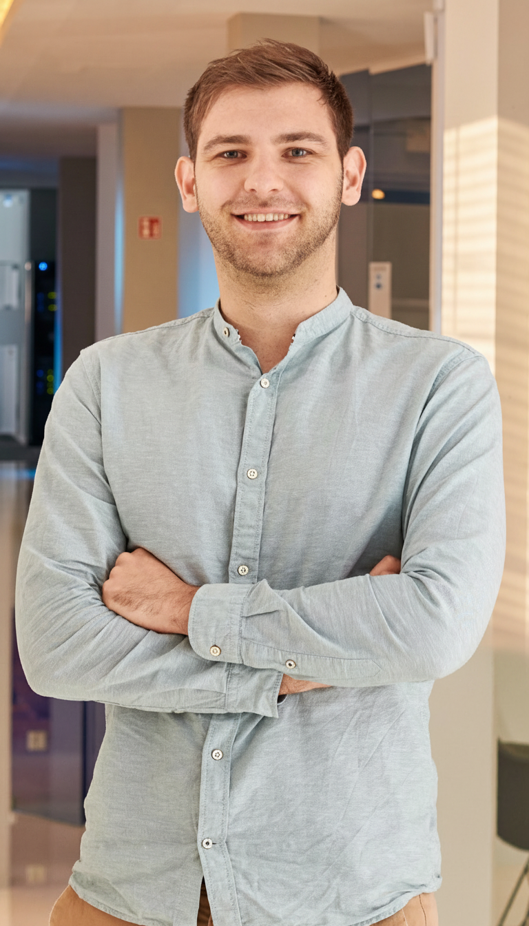 Ein männlicher Mann steht vor einem Hintergrund mit moderner Einrichtung, trägt eine hellblaue Hemd und lächelt.