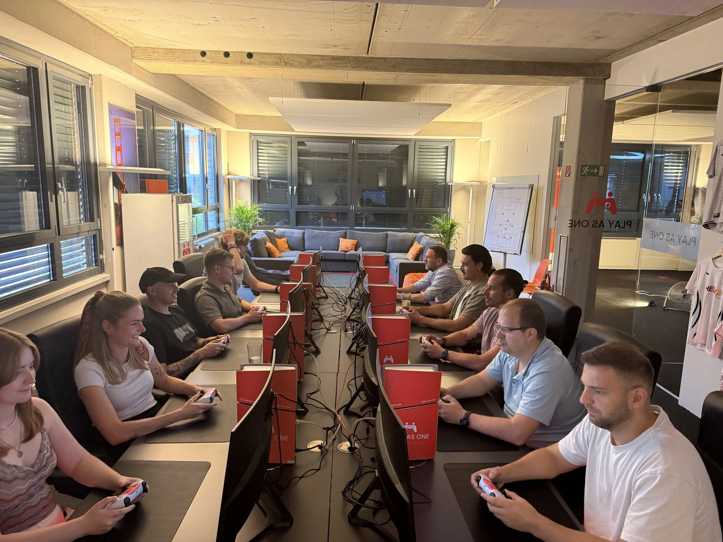 Mehrere Menschen spielen an Computertischen in einem modern eingerichteten Raum, vermutlich in einem Café oder Gemeinschaftsraum, mit großen Fenstern und Pflanzen im Hintergrund.