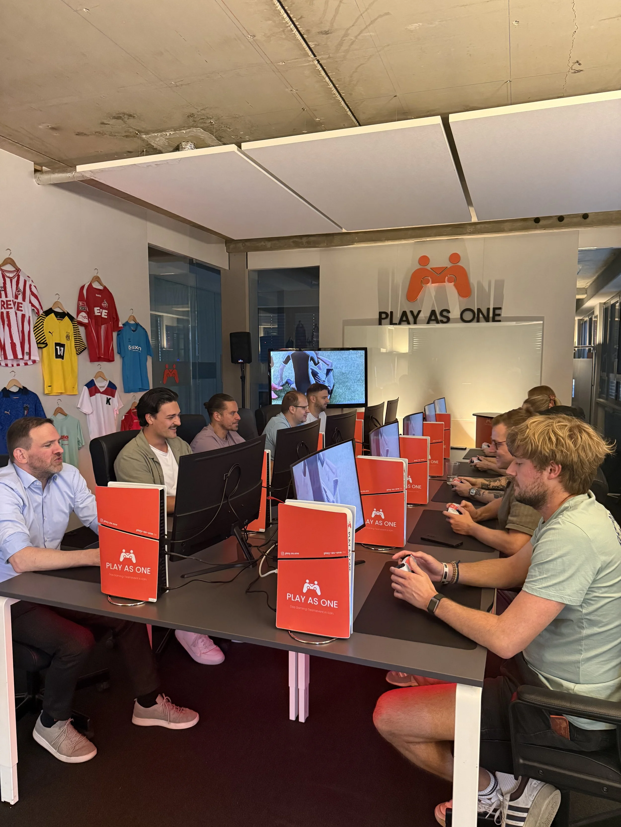 Menschen spielen Videospiele an einem langen Tisch in einem Raum mit dem Slogan "PLAY AS ONE" und roten Spiel-Controller-Symbolen auf Bildschirmen. An der Wand sind verschiedene sportliche Trikots hängt.