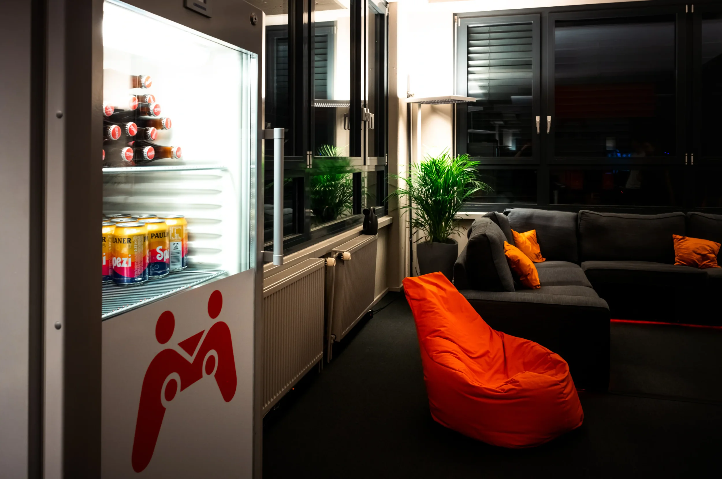 Ein Getränke-Kühlschrank mit Getränken, zwei grüne Pflanzen, ein schwarzes Sofa mit orangefarbenen Kissen, große Fenster mit geschlossen Rollläden, dunkles Zimmer, im Hintergrund eine Lampe.