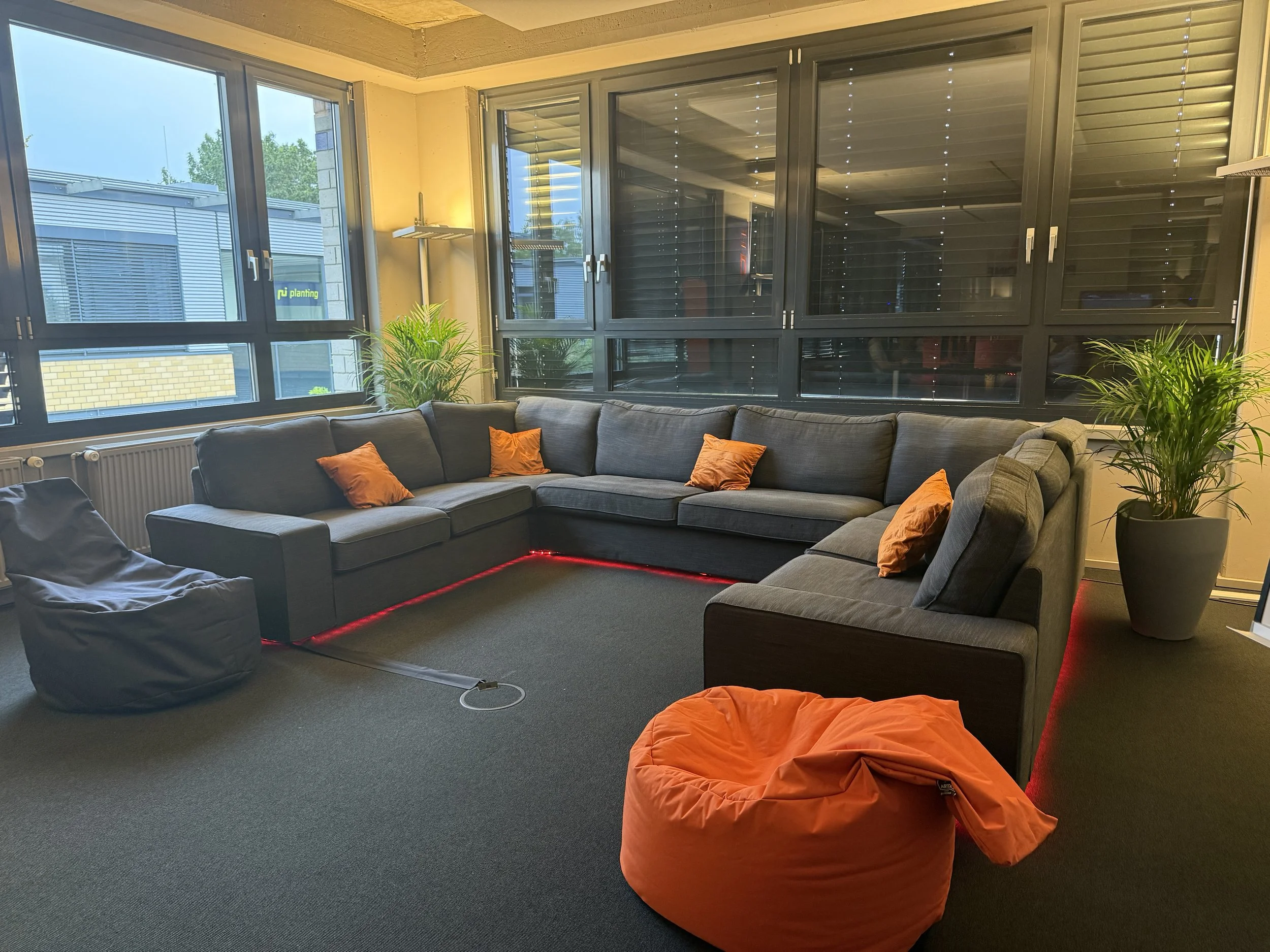 Wohnzimmer mit großen Fenstern, grauem Sofa mit orange Kissen, Pflanzen, Lampen, grauem Sessel und roter LED-Beleuchtung am Boden.