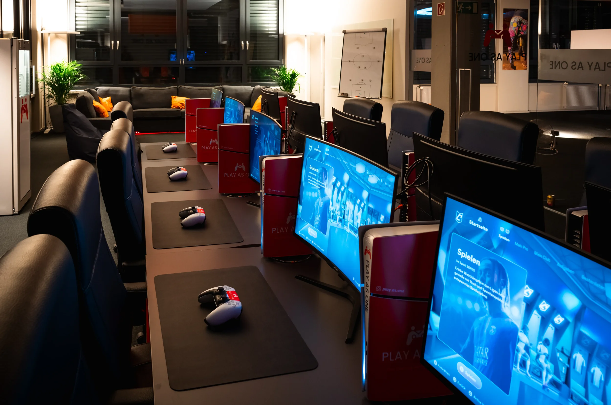 Ein Raum mit mehreren Computern, die Bildschirme zeigen, sowie einige Stühle, Pflanzen, eine Sofa-Ecke mit Kissen im Hintergrund, eine Whiteboard an der Wand und Glasfenster.