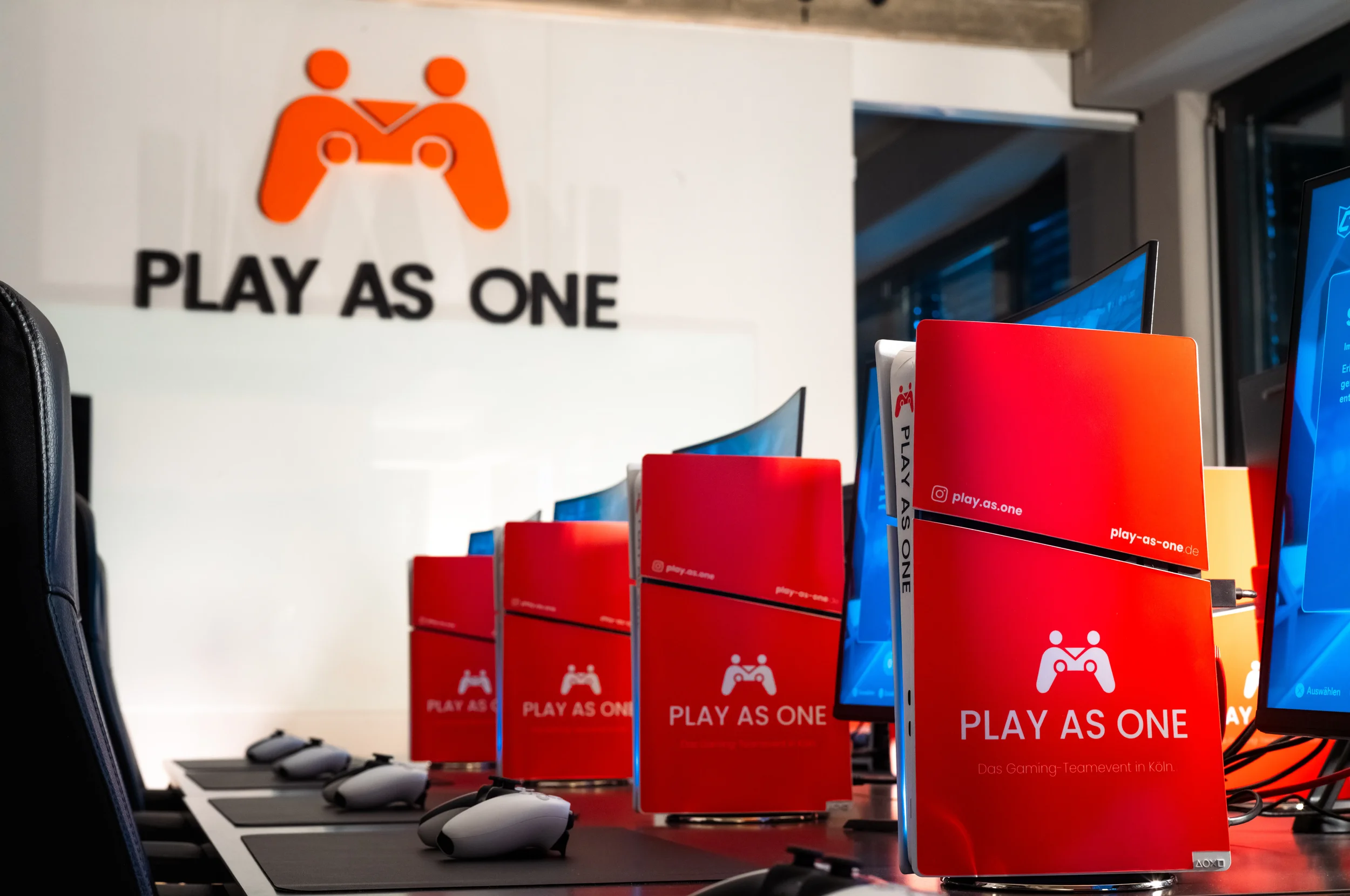Ein Gaming-Event in Köln mit mehreren Monitoren, rote Informationsstände mit dem Schriftzug 'PLAY AS ONE' und schwarzen Computermäusen auf Tischen.