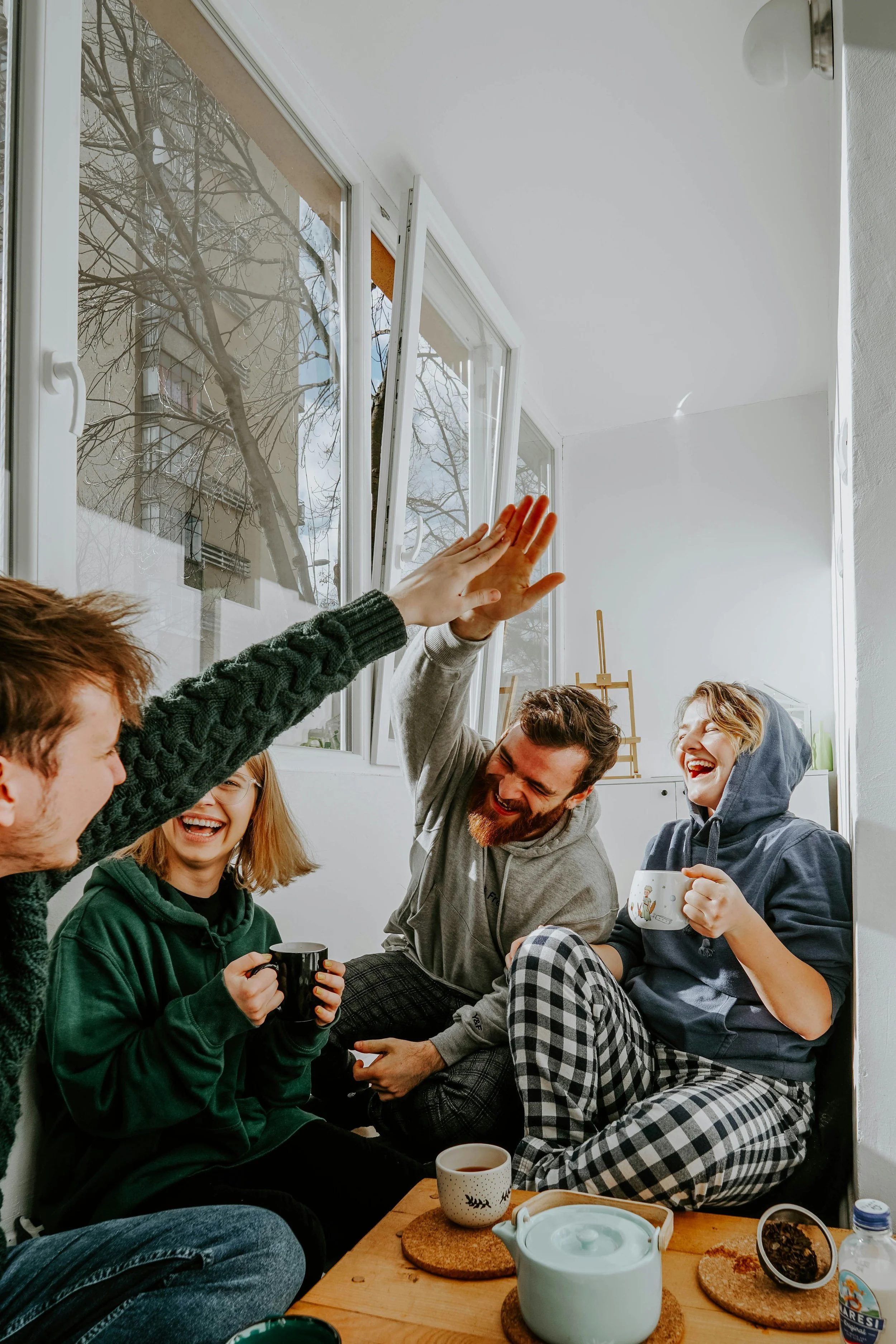 Eine Gruppe junger Menschen lachen und unterhalten sich bei einer Tasse Tee oder Kaffee in einem Raum mit großen Fenstern, durch die Bäume zu sehen sind.