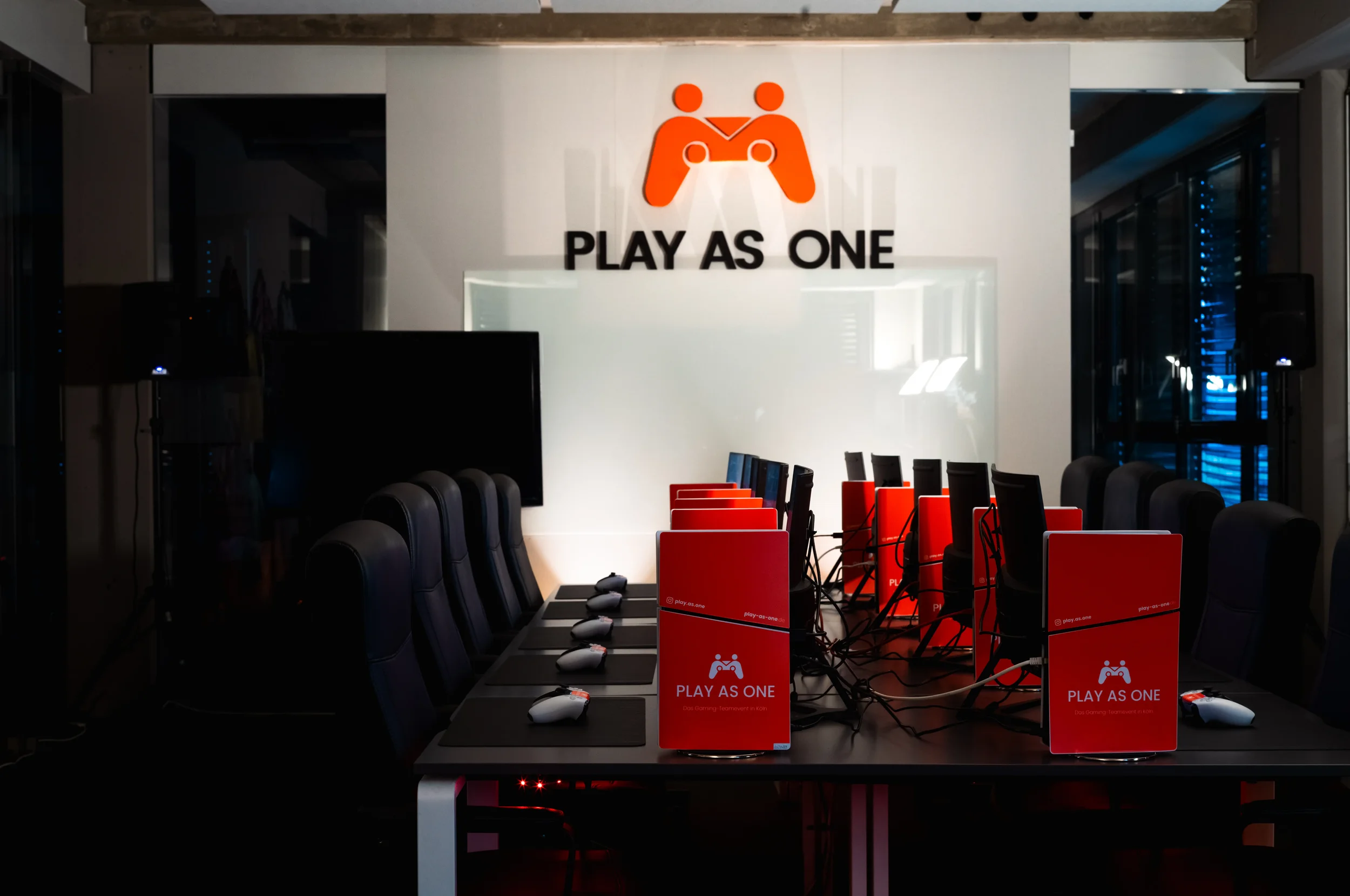 Ein Raum mit mehreren Gaming-Stationen, jeder mit einem Monitor, einer Gaming-Maus und einer Tastatur. Am oberen Ende des Bildes befindet sich ein großes Logo mit zwei orangefarbenen Figuren, die einander gegenüber sitzen und eine Spielkonsole halten