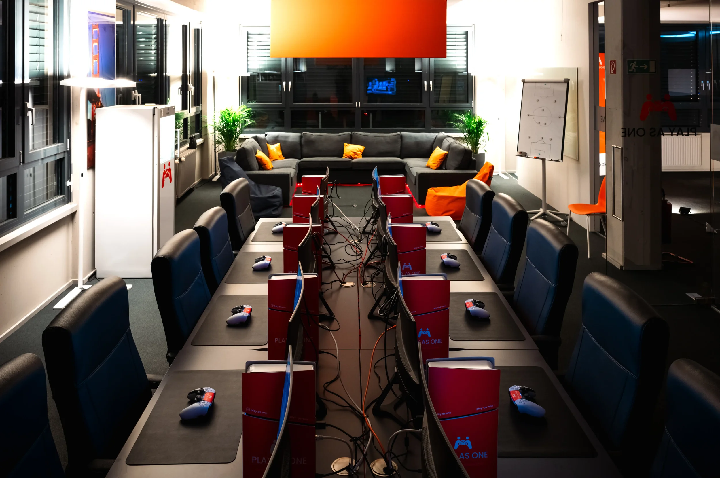 Ein moderner Raum mit mehreren Computertischen, einer großen sogenannten Gaming-Tafel, Sofas mit Orange Kissen, Pflanzen, und einem Flipchart, der für eine Gaming- oder Entertaining-Aktivität vorbereitet ist.
