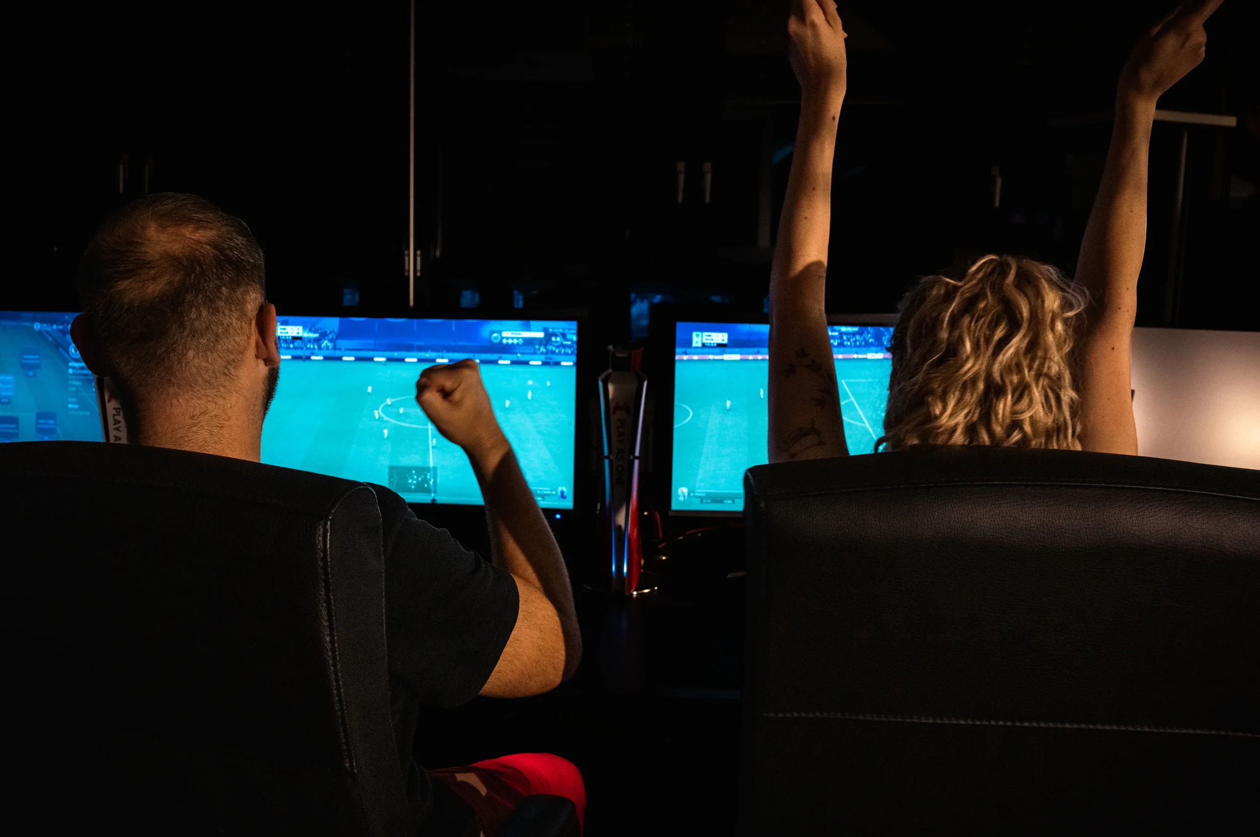 Zwei Menschen sitzen vor Bildschirmen und schauen ein Videospiel, ein Mann auf der linken Seite zeigt mit der Faust und ein Mädchen auf der rechten Seite hebt die Arme in der Luft.