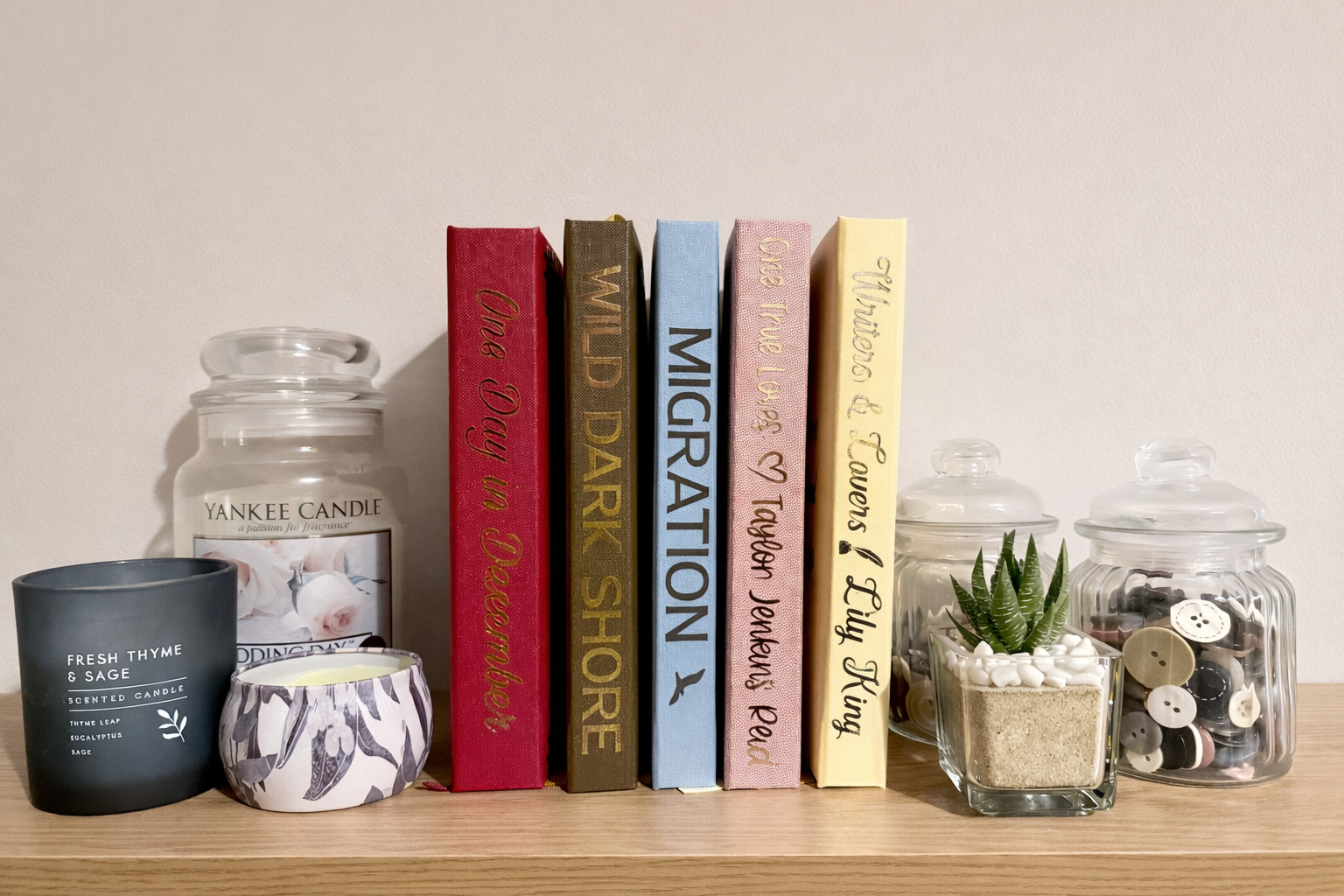 Shelf with candles, decorative bespoke custom rebound books, and storage jars, including a black candle labelled 'Fresh Thyme & Sage,' a large Yankee Candle jar, and smaller jars with buttons and a plant.
