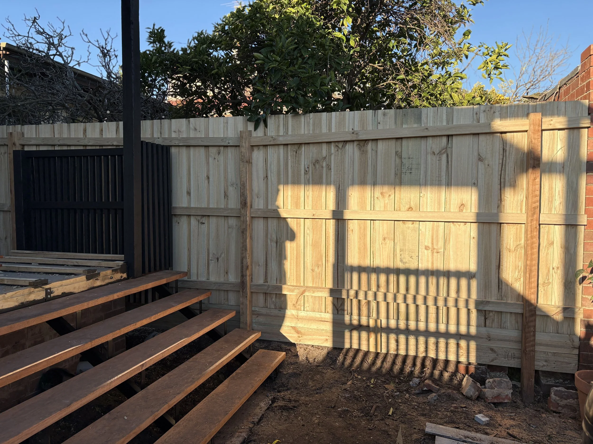 A backyard scene showing a newly installed wood fence with sunlight casting shadows of a staircase railing. The area is bare soil with some bricks and construction debris, and a wooden staircase is partially built on the left side.
