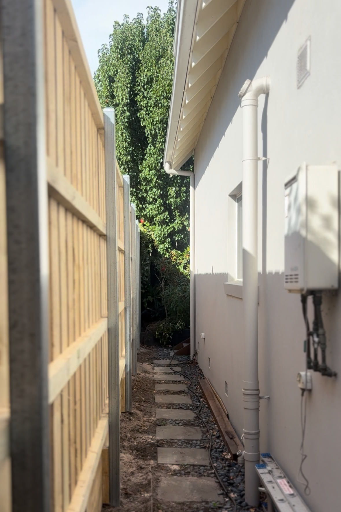 Narrow backyard pathway with stepping stones, fenced on the left and a white house wall with pipes and electrical boxes on the right, and greenery in the background.