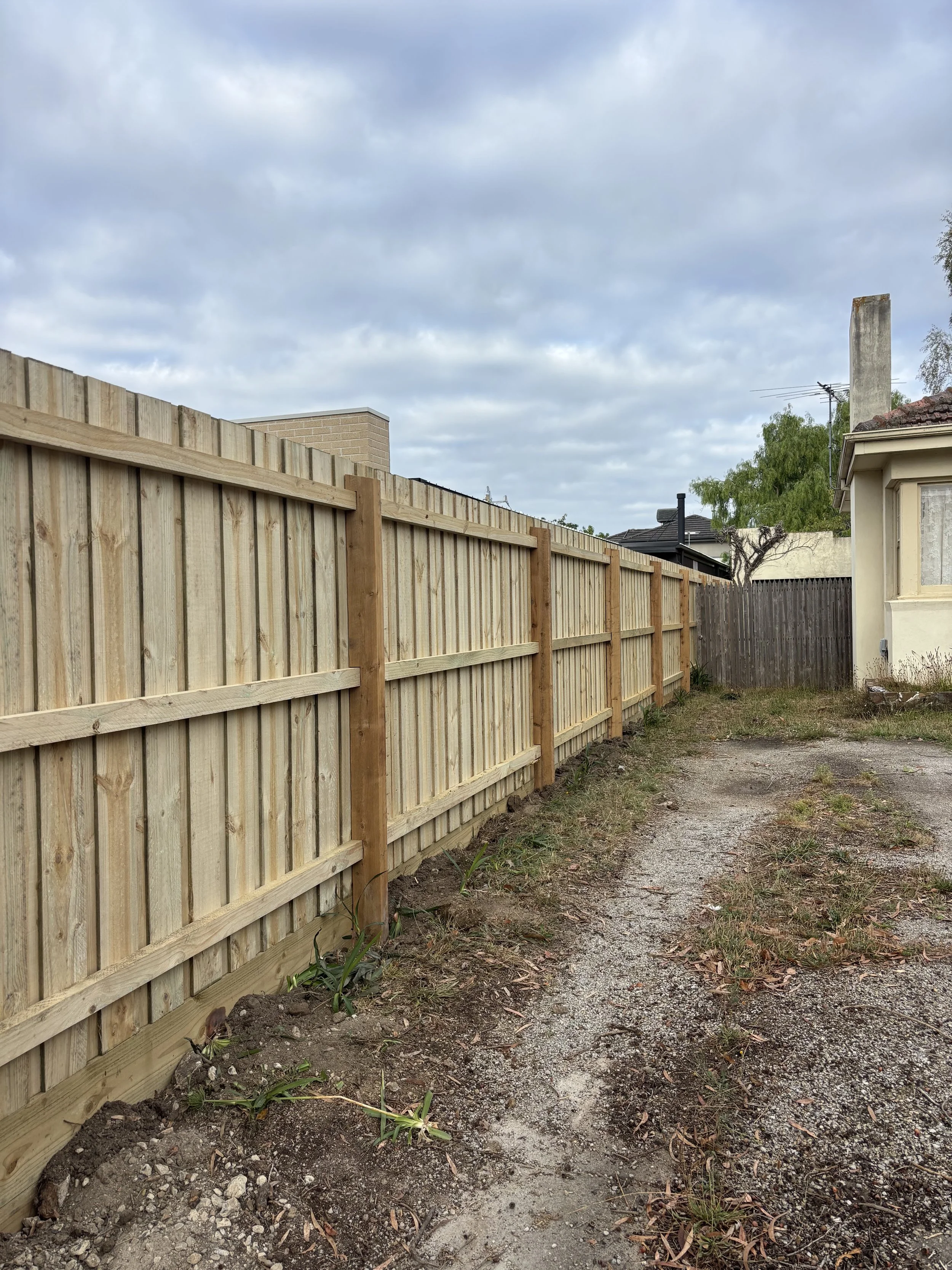 A new wooden fence along a dirt driveway outside a house under a cloudy sky.